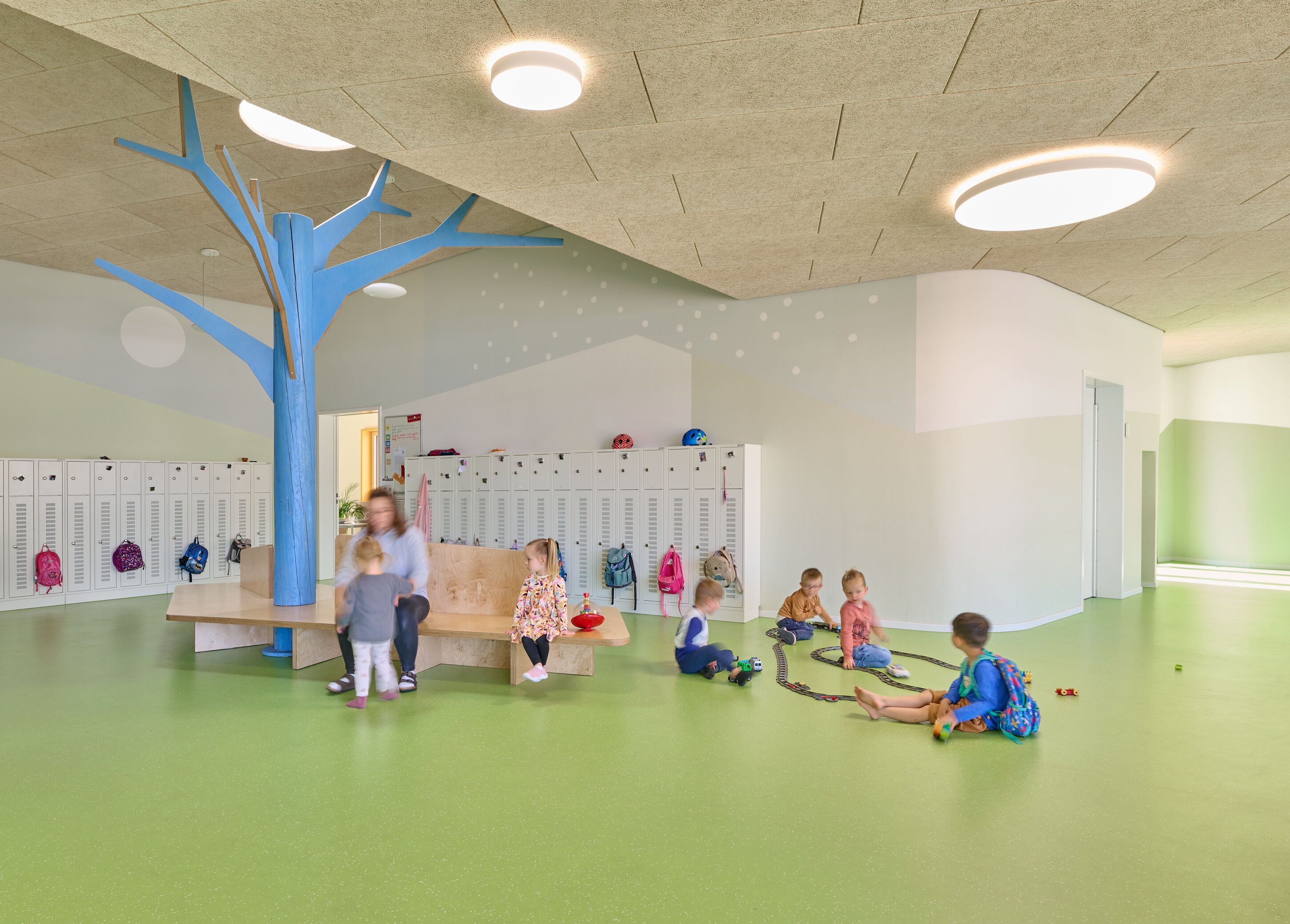 Project by Behnisch Architekturbuero, KiTa Pioneer-Kaserne. Children playing on a green floor in a daycare room with lockers, a bench, and a blue tree-like column, while an adult sits nearby.