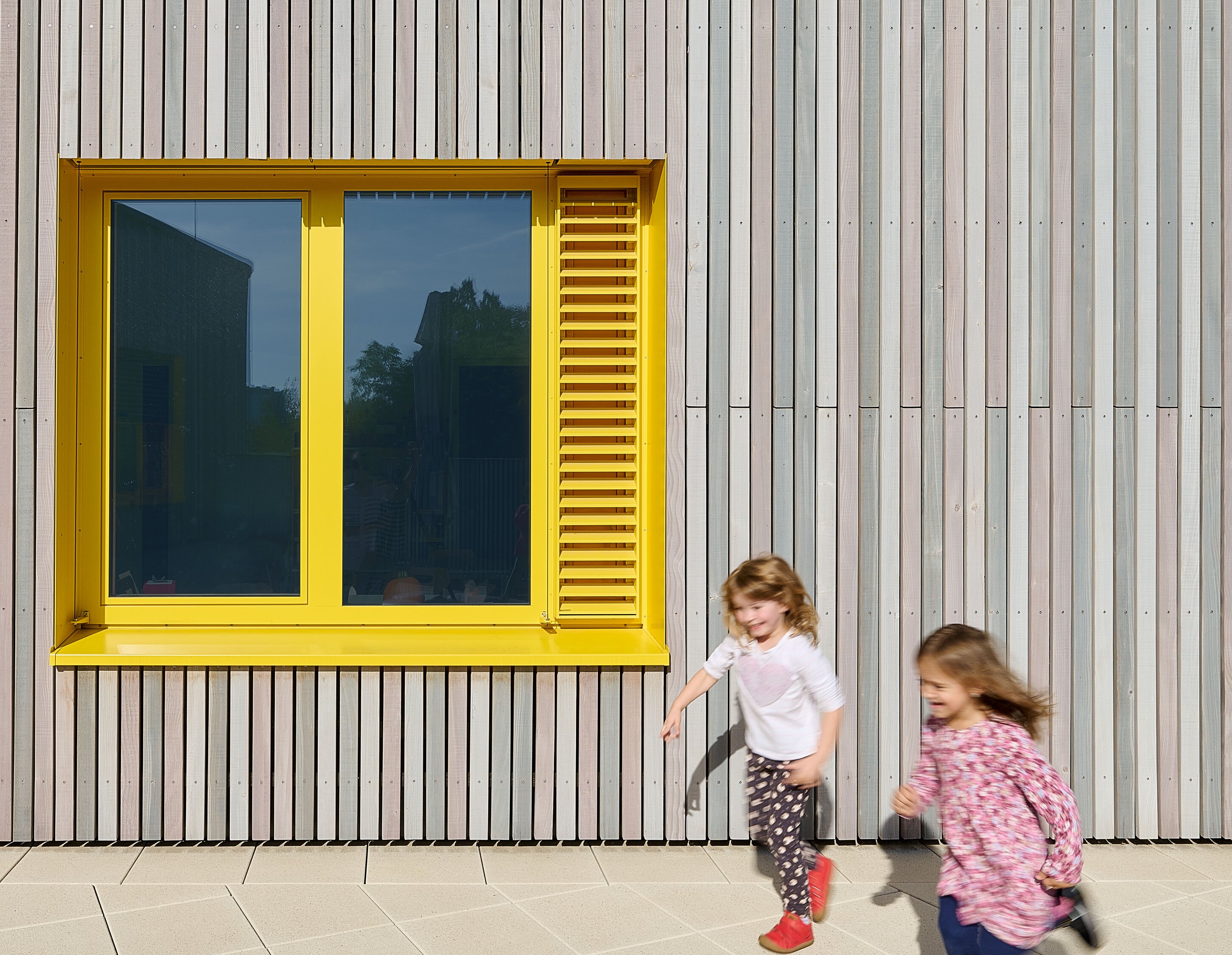 Project by Behnisch Architekturbuero, KiTa Pioneer-Kaserne. Two children running past a building facade with vertical cladding and a bright yellow-framed window.