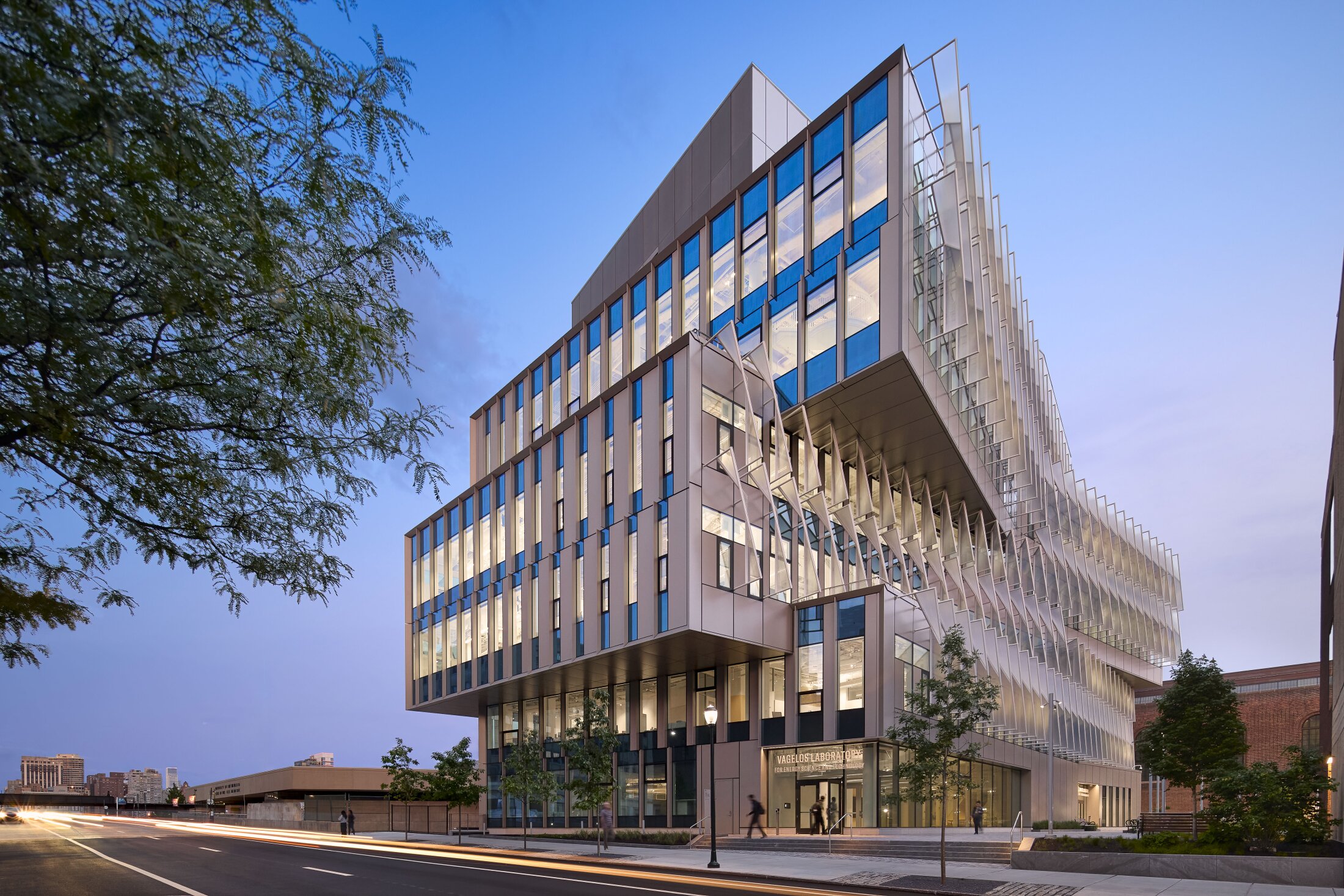 Project by Behnisch Architekturbuero, The Vagelos Laboratory for Energy Science & Technology, University of Pennsylvania. Modern office building with glass facade and vertical fins, illuminated at dusk in city street. On the right side of the building there is the sun protection.