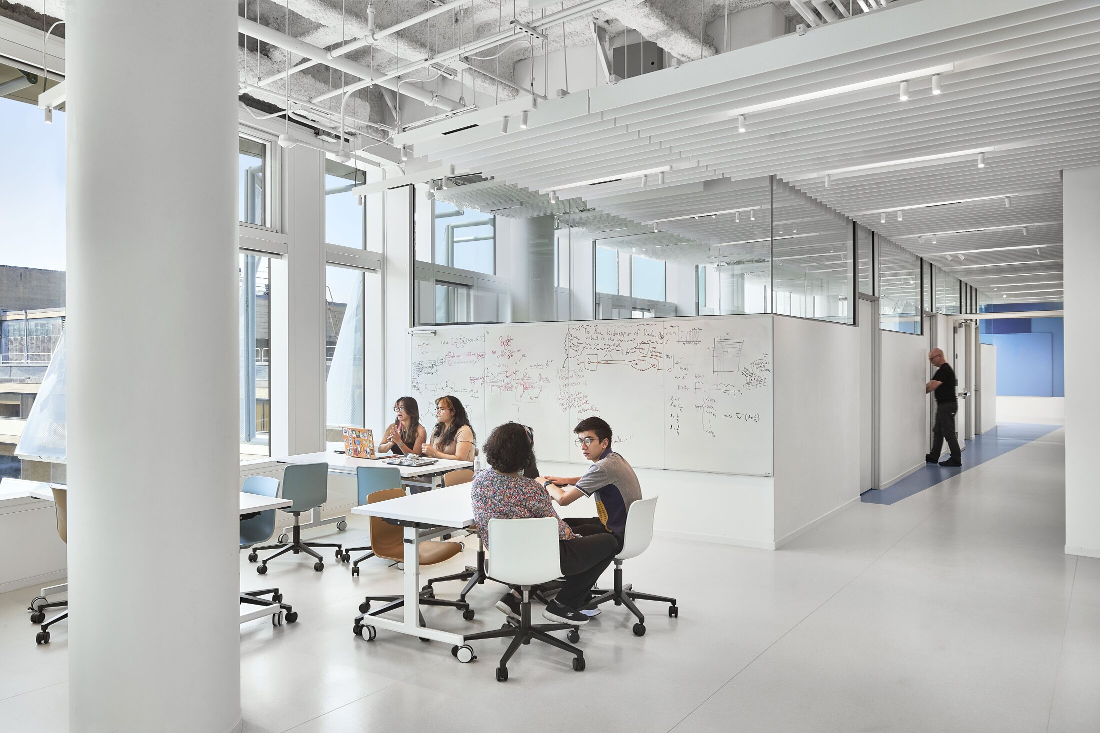 Project by Behnisch Architekturbuero, The Vagelos Laboratory for Energy Science & Technology, University of Pennsylvania. Students are working at tables in a bright studio with large windows and whiteboard drawings. In the background, a corridor can be seen leading to other areas of the building.