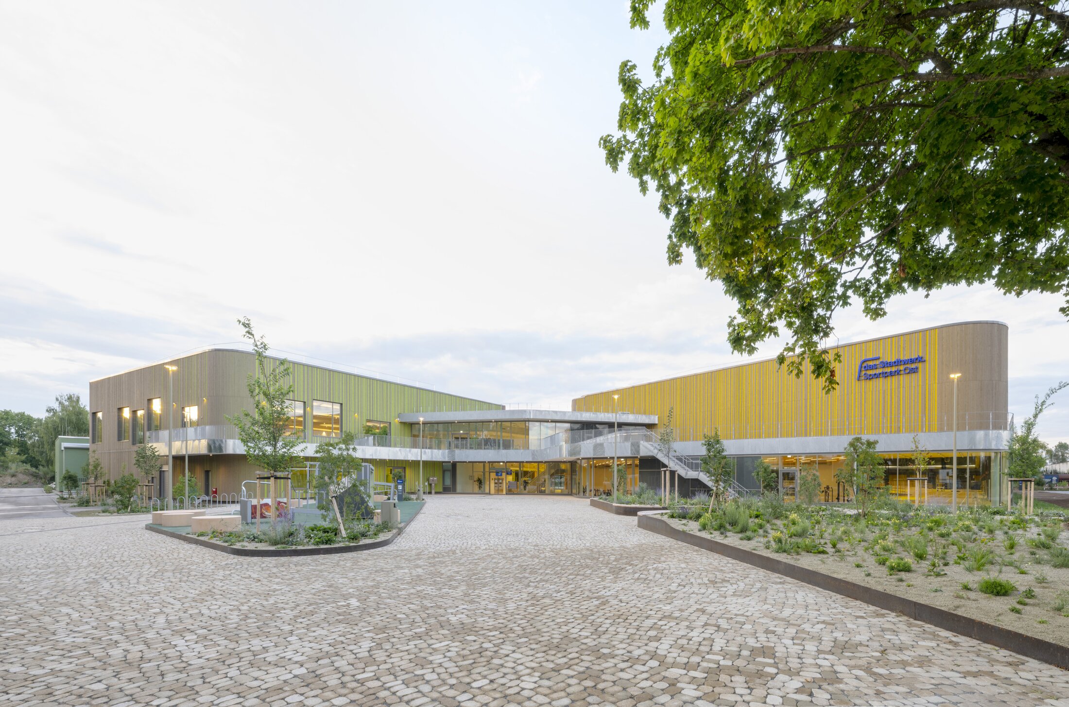 Project by Behnisch Architekturbuero, Max Aicher Arena. Mountain valley with forested slopes, a village of houses and a church steeple, the white-roofed building in the distance.