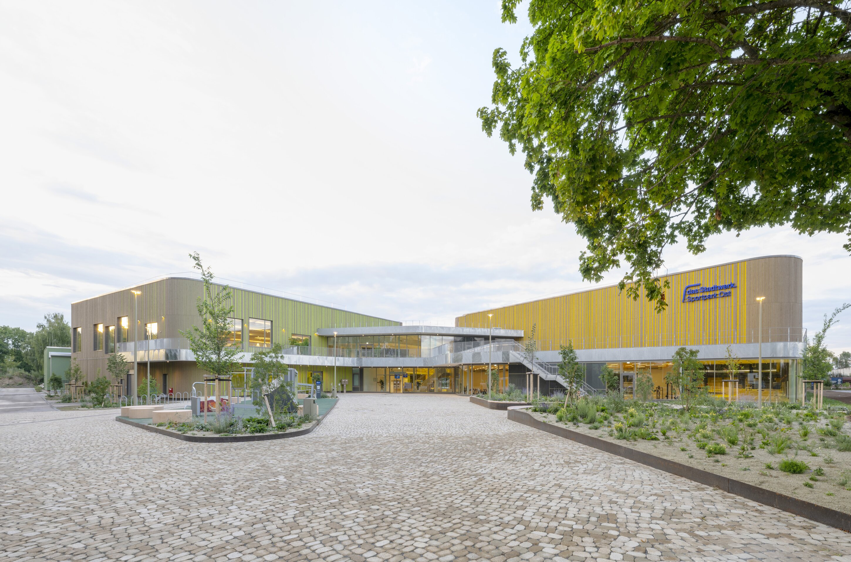Project by Behnisch Architekturbuero, New Construction of an Athletics Training Hall and an Indoor Swimming Pool. Modern sports complex with green and yellow facades, glass entrance, and landscaped courtyard with trees and cobblestone paving.