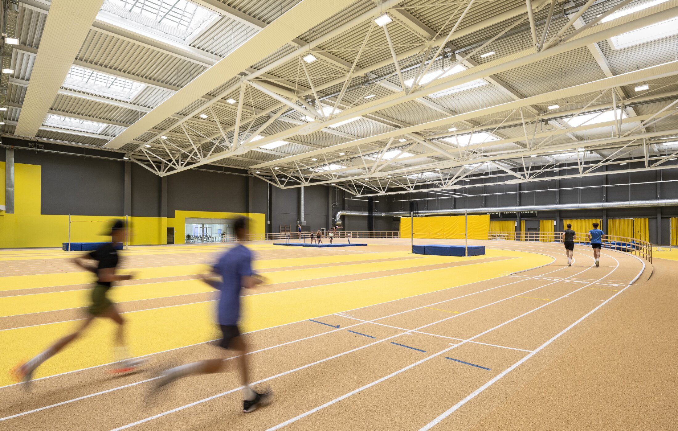 Project by Behnisch Architekturbuero, Sportpark Ost Regensburg. Athletics hall with runners on the tartan track. The floor is partially yellow. The interior has high ceilings with skylights and several yellow accents.