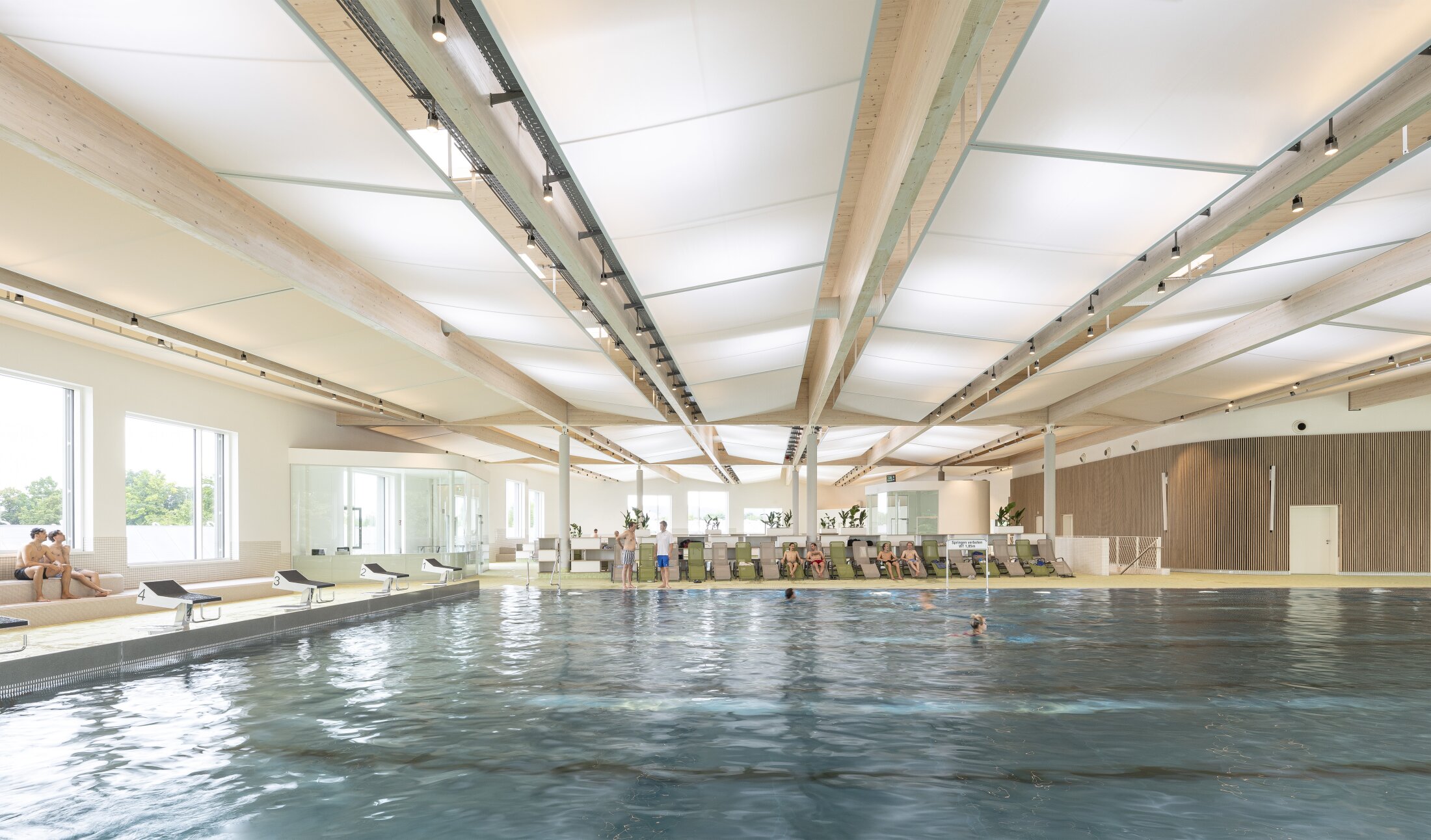 Project by Behnisch Architekturbuero, Sportpark Ost Regensburg. Indoor swimming pool with a seating area for visitors, large windows, and a wooden beamed ceiling. People are swimming in the large pool. Starting blocks can be seen on the left side of the pool.