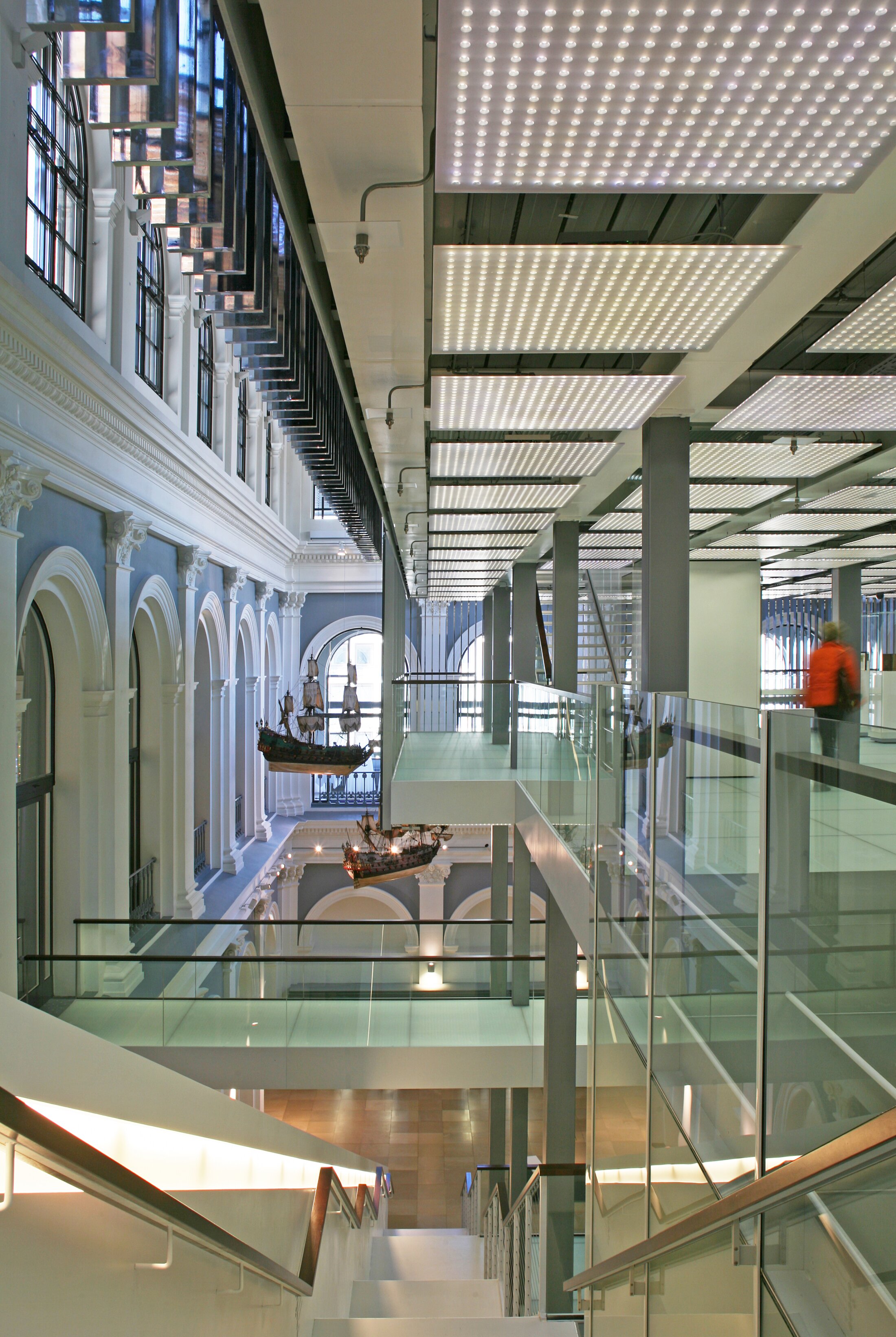 Project by Behnisch Architekturbuero, Haus im Haus. Interior view of a historic hall with a modern glass walkway, stairs, LED ceiling panels, and hanging ship models visible below.
