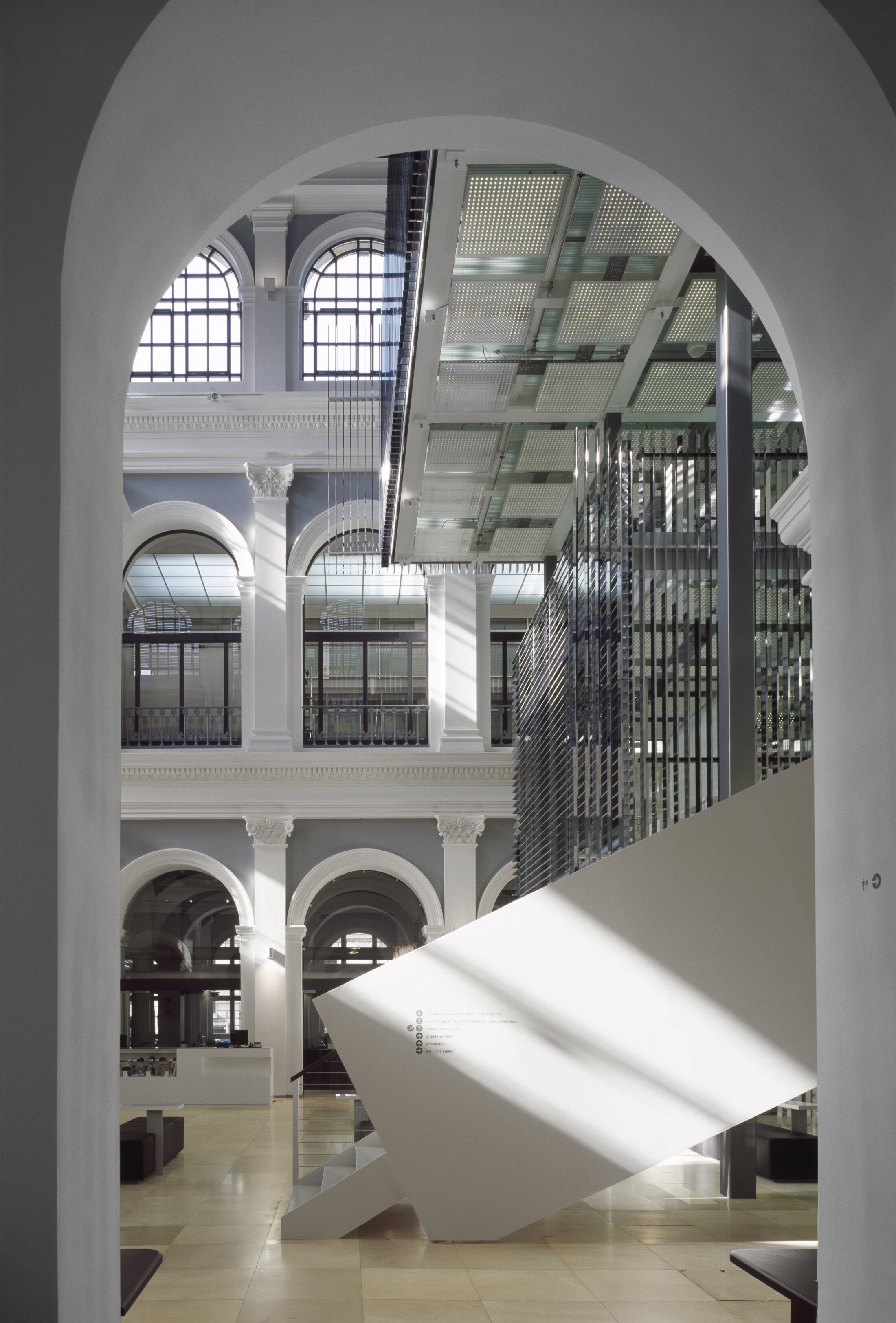 Project by Behnisch Architekturbuero, Haus im Haus. View through an archway into a historic hall with columns, arched windows, and a modern glass ceiling and angular white staircase.