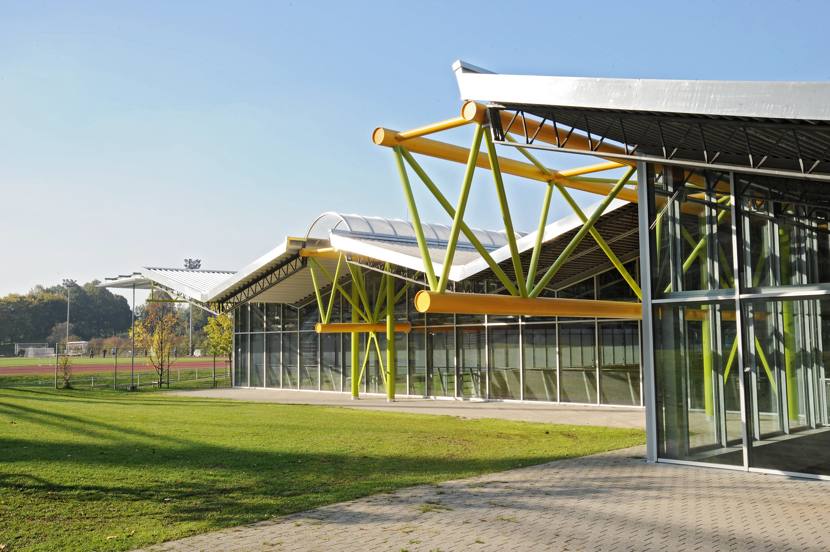 Project by Behnisch Architekturbuero, Werner-von-Linde-Halle. Modern sports facility with glass facade, sloping roofs and green and yellow steel beams, next to a grass field surrounded by a tartan track.