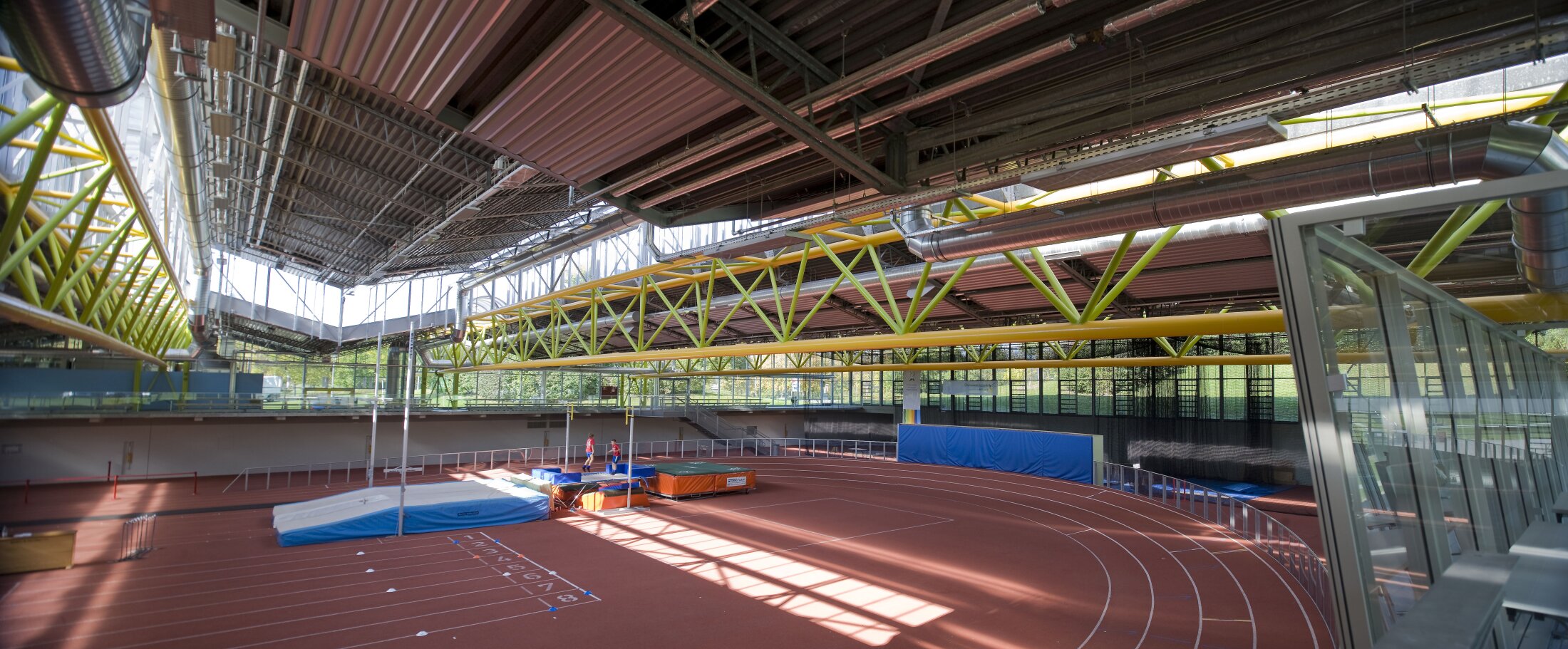 Project by Behnisch Architekturbuero, Werner-von-Linde-Halle. Athletics hall with tartan track, high jump mats and exposed steel beams and pipes under a large glass roof.
