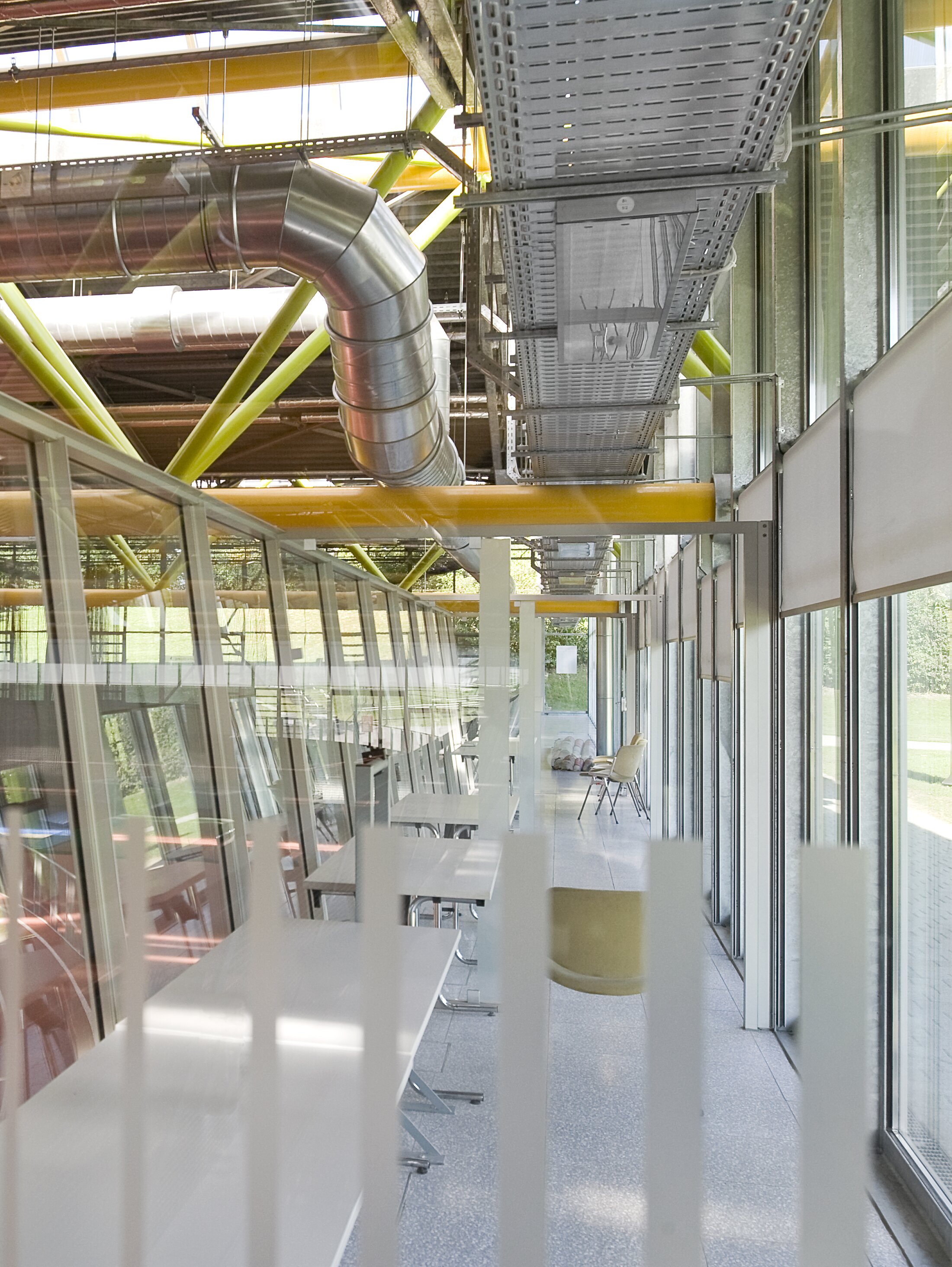 Project by Behnisch Architekturbuero, Werner-von-Linde-Halle. Glass-walled corridor with tables and chairs, exposed ducts and yellow beams overhead, overlooking the indoor sports track.