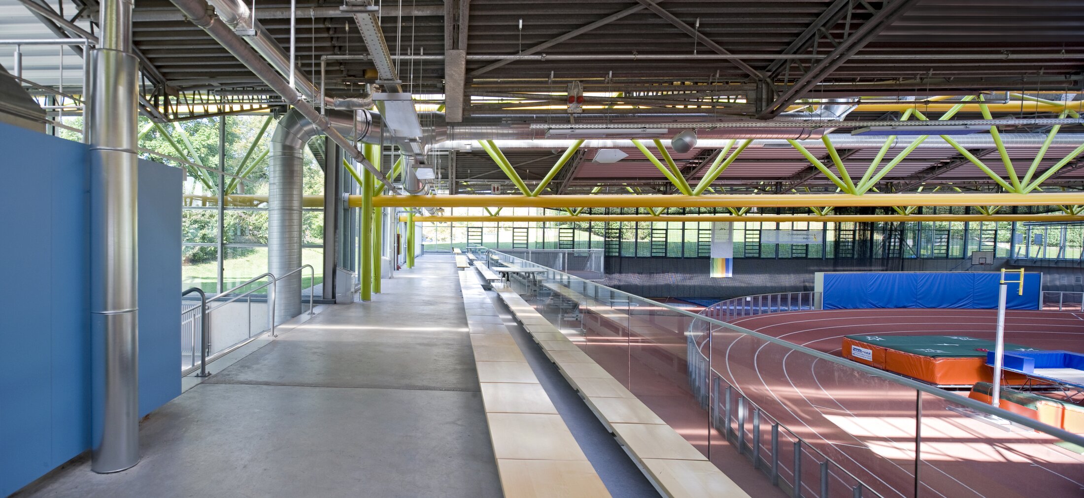 Project by Behnisch Architekturbuero, Werner-von-Linde-Halle. Athletics hall with a tartan track and high jump mats. Above, a spectator stand with seating. Above, exposed pipes and green steel girders under a large roof.