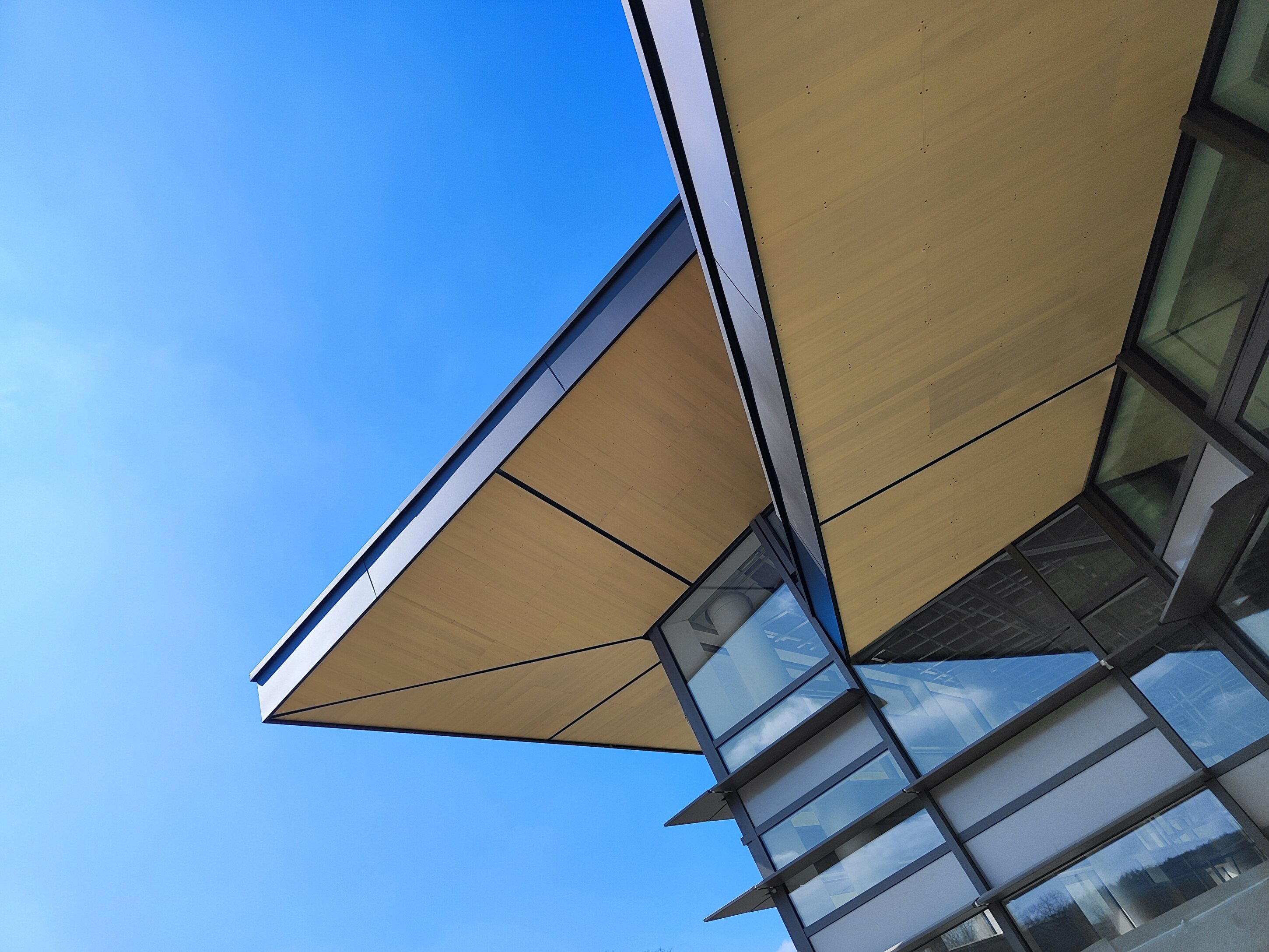 Project by Behnisch Architekturbuero, Bavarian Police Eichstaett. Upward view of a modern building corner with glass walls and a projecting roof, showing wood soffit panels against a clear blue sky. Project by Behnisch Architekturbuero, Bavarian Police Eichstaett.