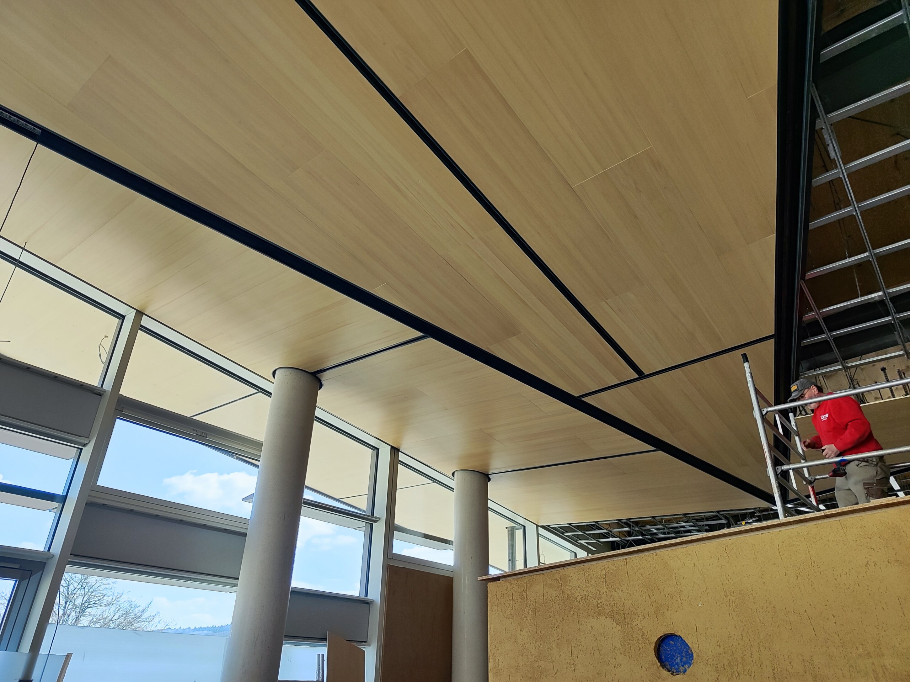 Project by Behnisch Architekturbuero, Bavarian Police Eichstaett. Interior under construction with wood ceiling panels, large windows, columns, and a worker on scaffolding installing ceiling elements.