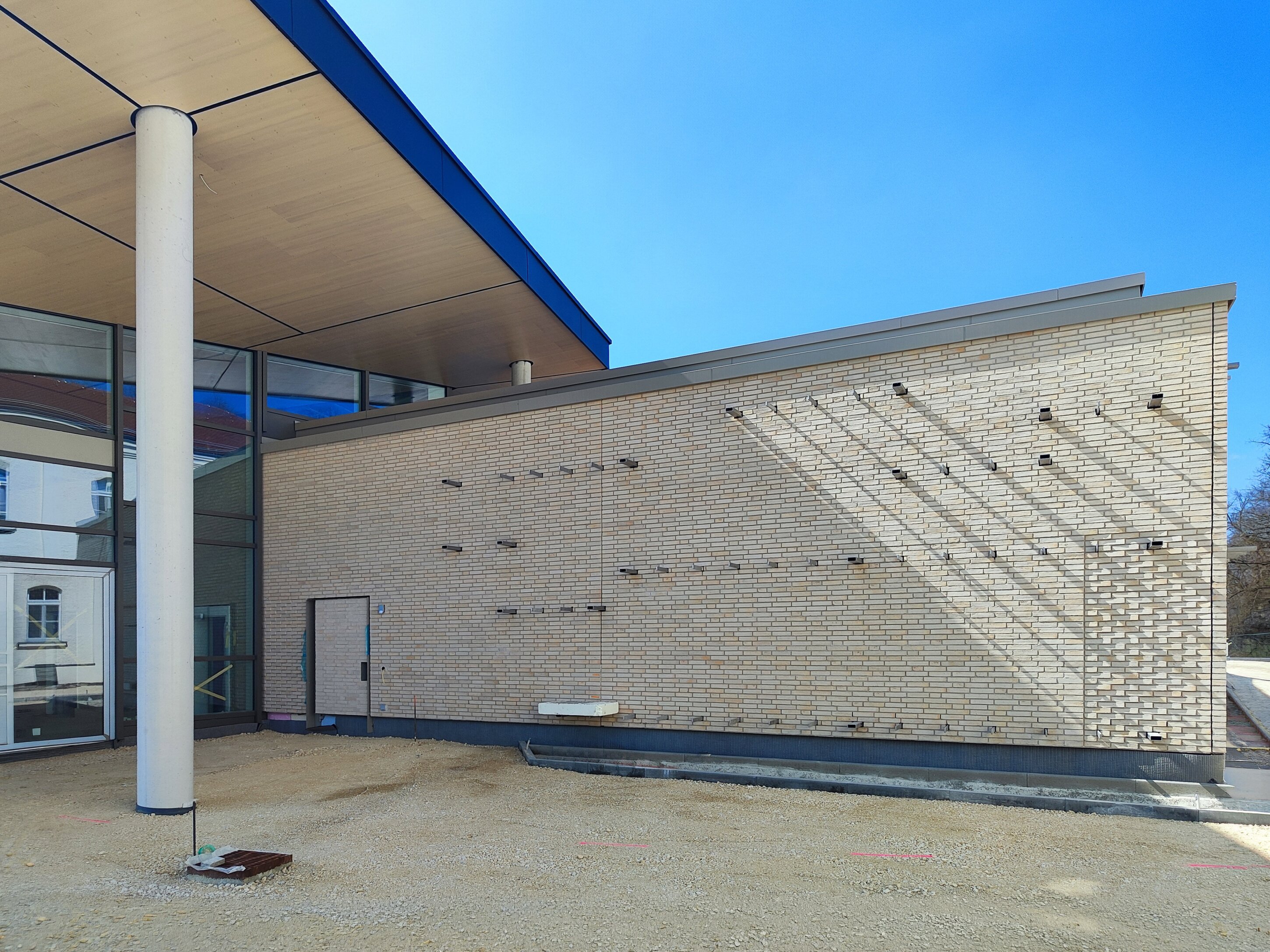 Project by Behnisch Architekturbuero, Bavarian Police Eichstaett. Exterior view of a modern building corner with brick wall, glass facade, and overhanging roof; sunlight casts shadows on the wall.
