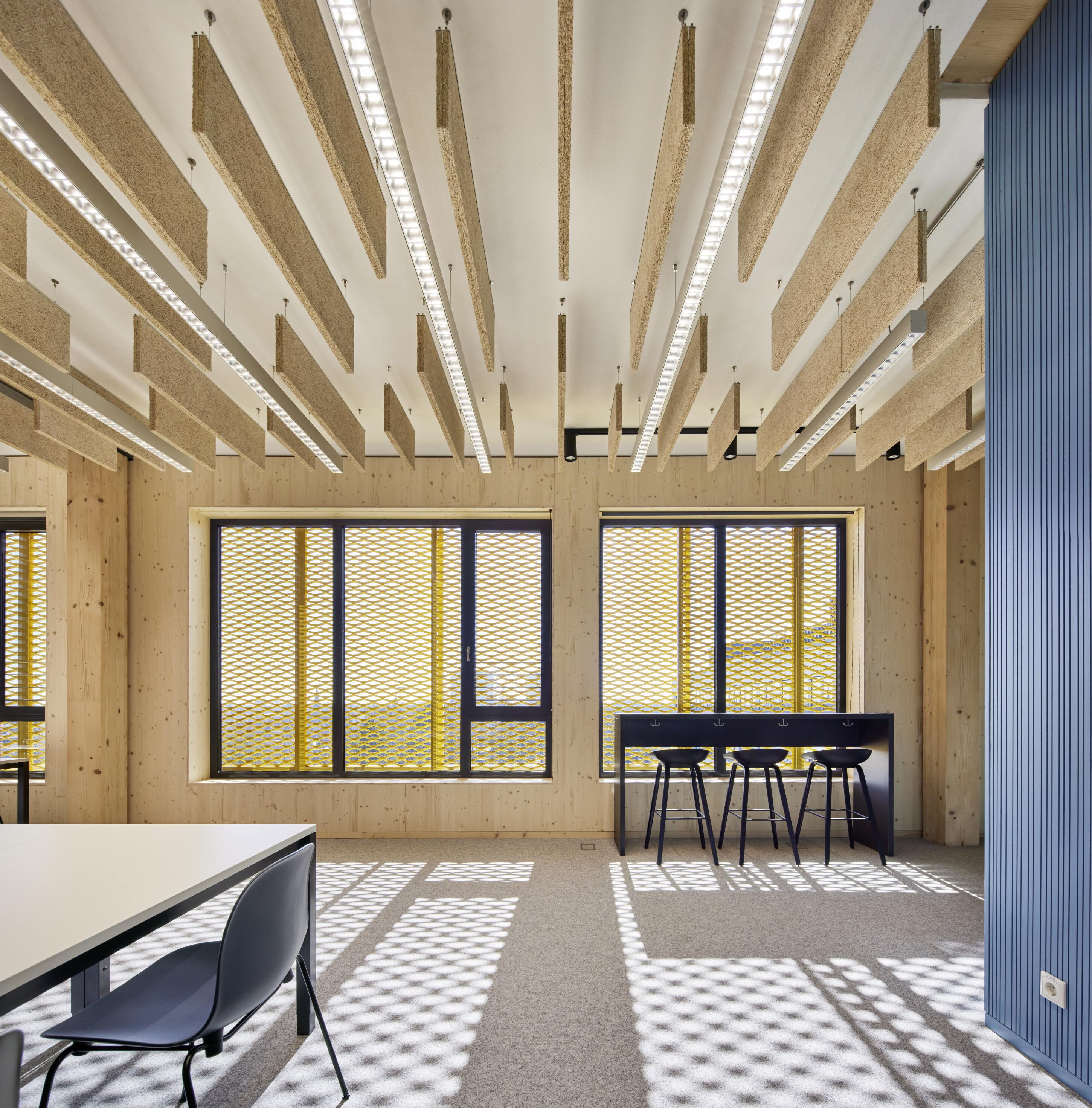 Project by Behnisch Architekturbuero, Extension Fachhochschulzentrum Münster University of Applied Sciences. Room with raised desks and stools in front of a large window. On the left hand side there is a Desk with chairs in the middle of the room and suspended acoustic ceiling panels.