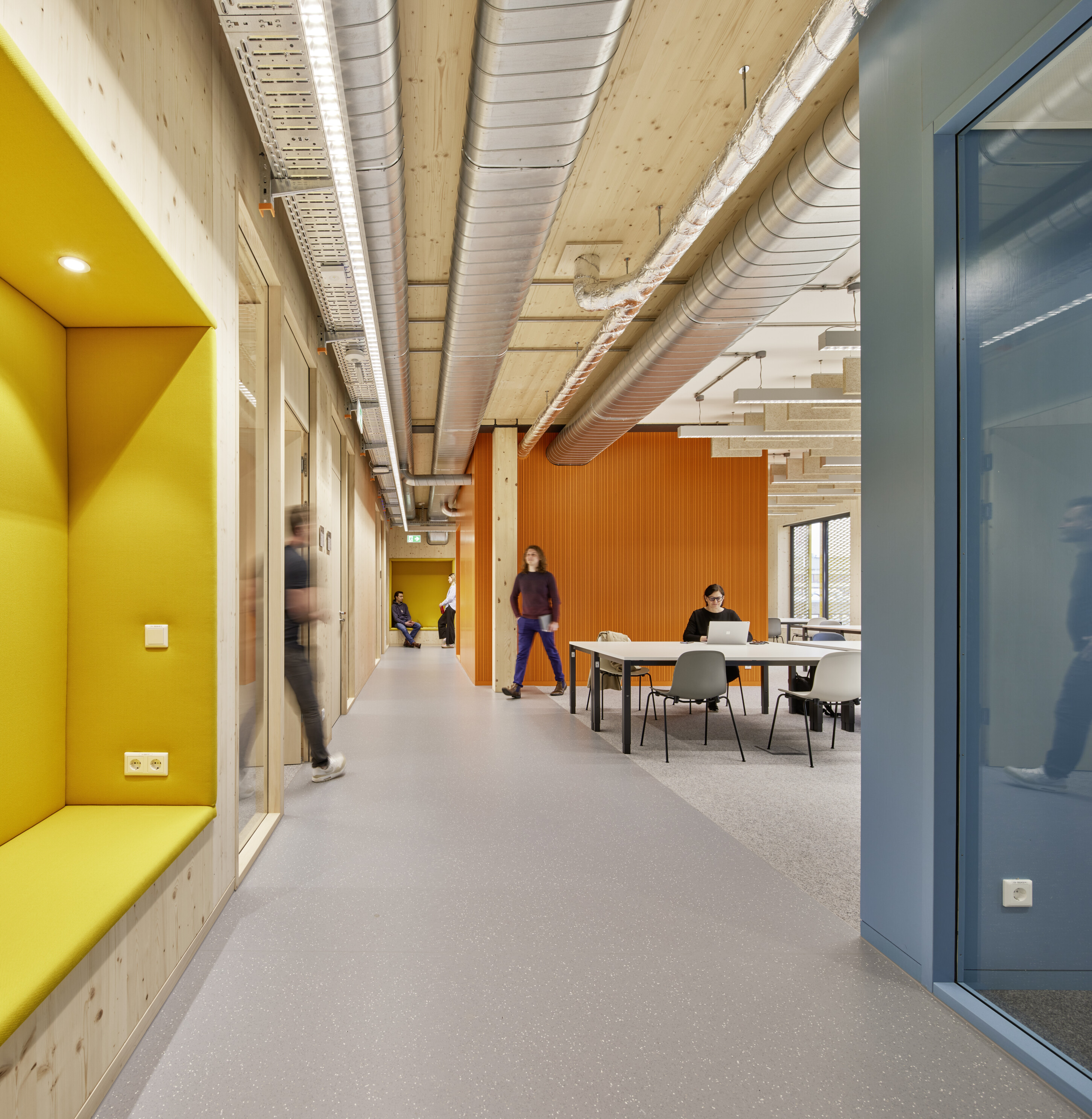 Project by Behnisch Architekturbuero, Extension Fachhochschulzentrum Münster University of Applied Sciences. Interior hallway with exposed ducts and wood ceiling, bright yellow and orange walls, and people walking or working at tables.