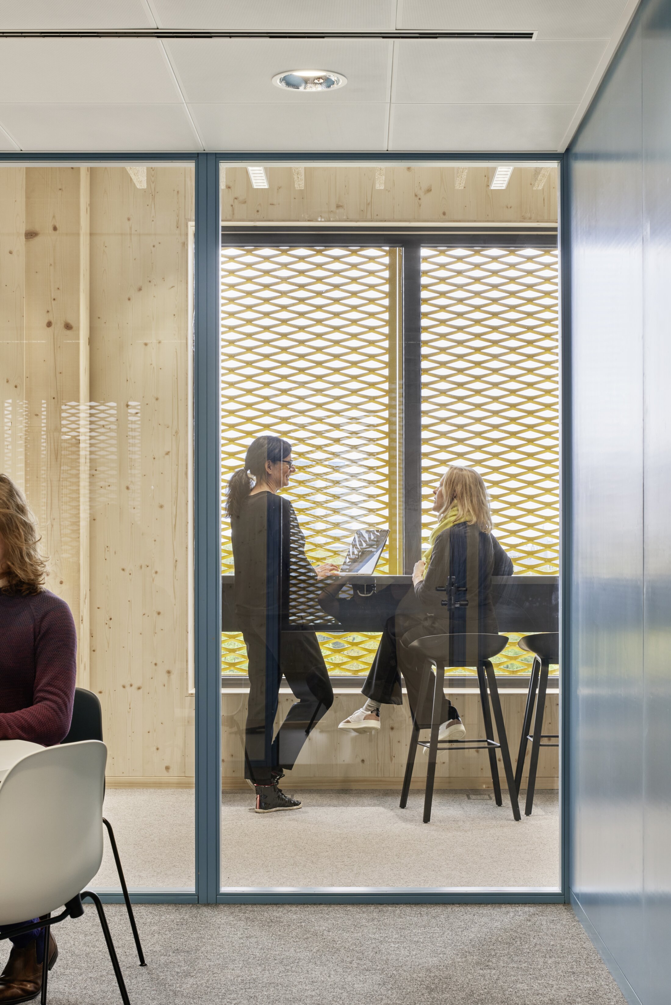 Project by Behnisch Architekturbuero, Extension Fachhochschulzentrum M&uuml;nster University of Applied Sciences. Two people are talking at a standing table in front of a window. The scene was captured through a glass partition in a meeting room.
