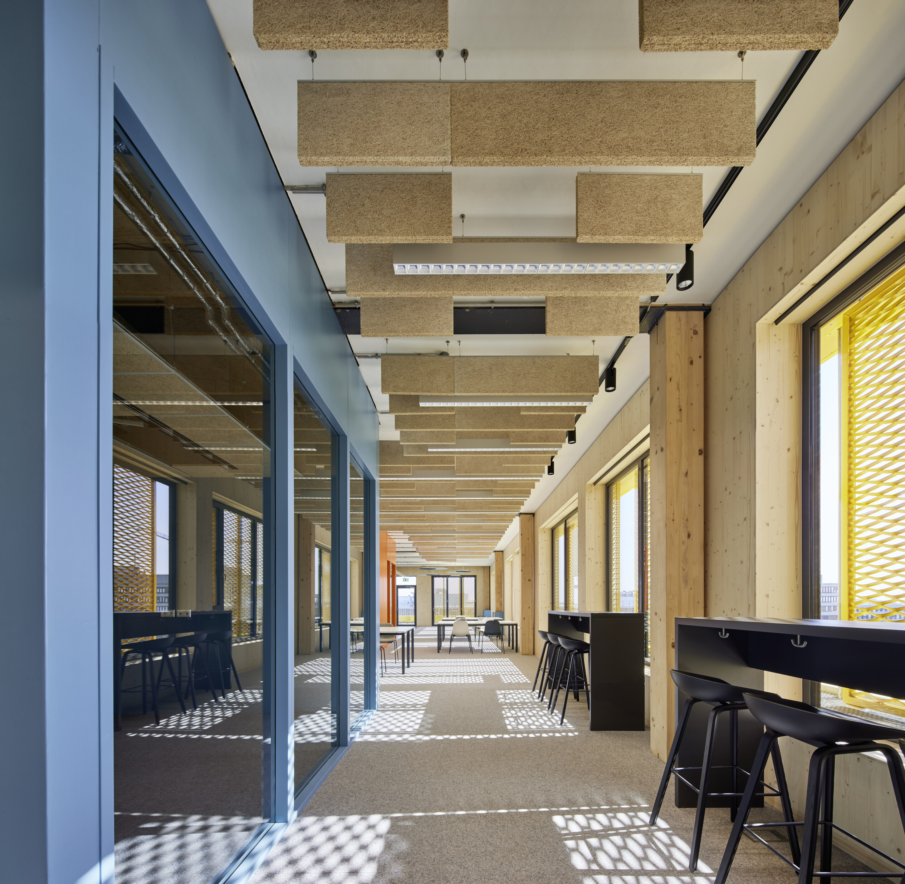 Project by Behnisch Architekturbuero, Extension Fachhochschulzentrum Münster University of Applied Sciences. Hallway with raised desks and stools, large windows and suspended acoustic ceiling panels.
