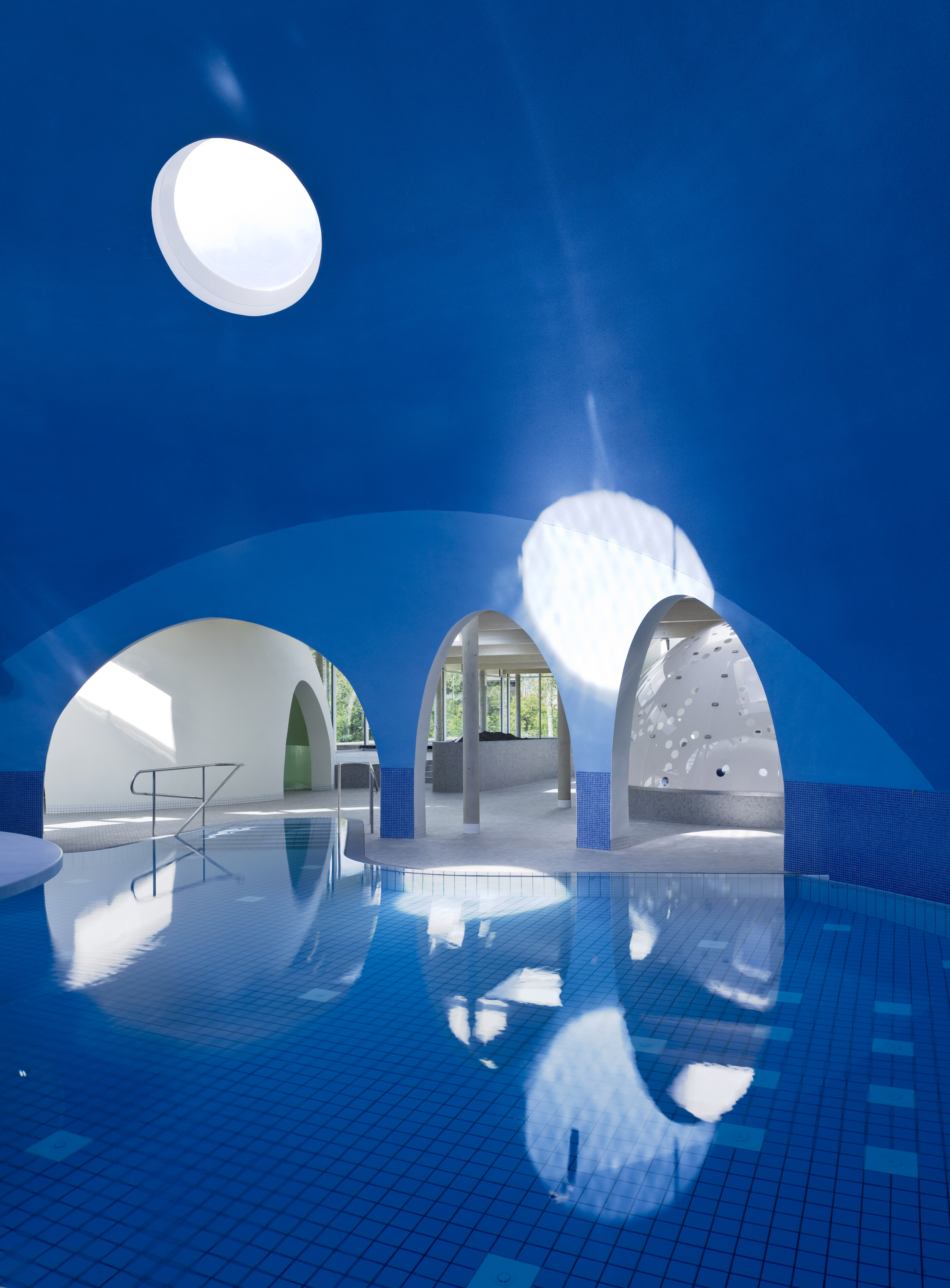 Project by Behnisch Architekturbuero, Thermal Spa Bad Aibling. Indoor pool beneath a blue domed ceiling with circular skylight and white arched openings, with reflections on the water surface.