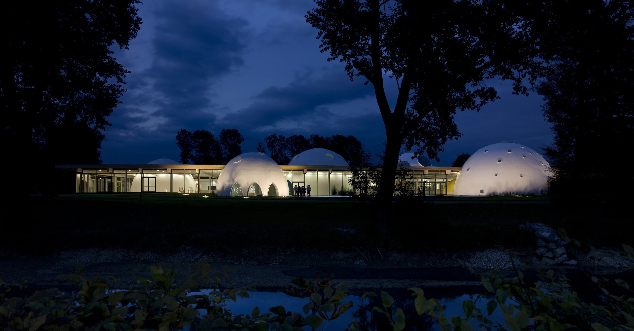 Project by Behnisch Architekturbuero, Thermal Spa Bad Aibling. Illuminated, dome-shaped buildings, connected at night by a building with a glass facade, nestled in a landscape with trees and a reflecting stream.