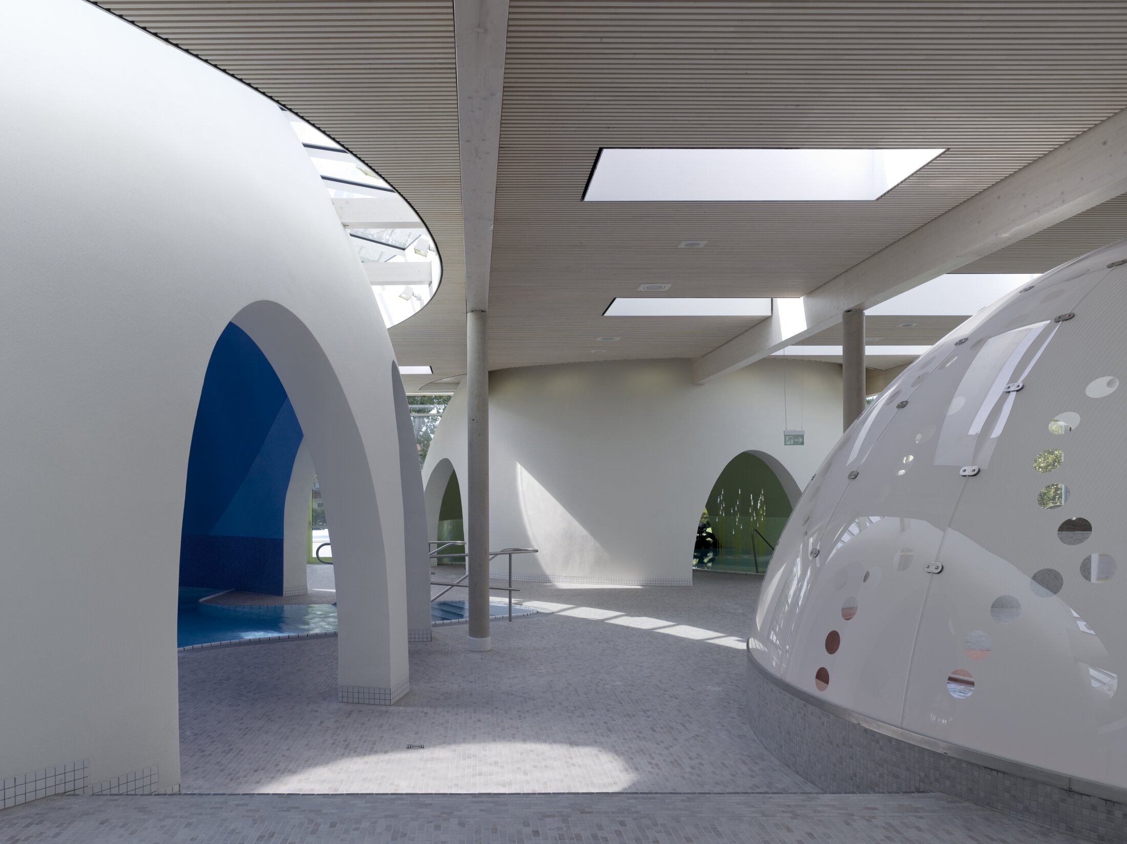 Project by Behnisch Architekturbuero, Thermal Spa Bad Aibling. Indoor pool area with white curved structures, arched openings, skylights, and tiled floors, with small pools visible beneath the arches.