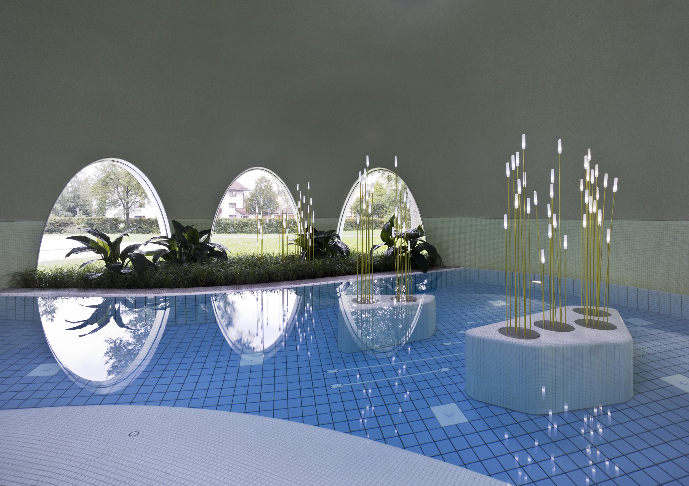 Project by Behnisch Architekturbuero, Thermal Spa Bad Aibling. Indoor pool with blue tiles, oval windows, indoor plants, and sculptural light features rising from platforms in the water.