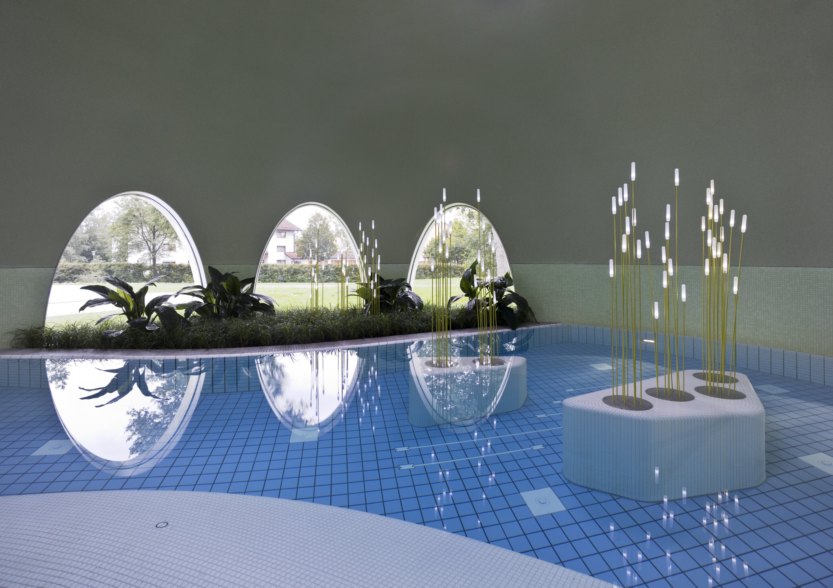 Project by Behnisch Architekturbuero, Thermal Spa Bad Aibling. Indoor pool with blue tiles, oval windows, indoor plants, and sculptural light features rising from platforms in the water.