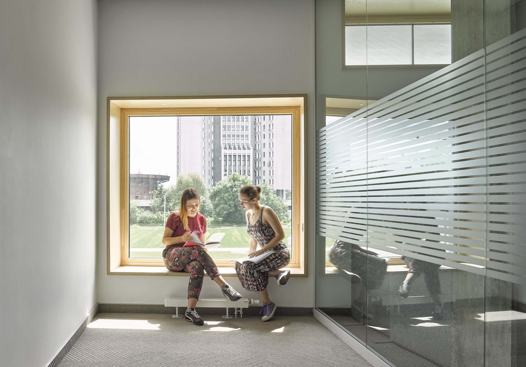 Project by Behnisch Architekturbuero, The Metropolitan Sheptytsky Center, Ukrainian Catholic University Lviv. Two people sit on a window bench reading and talking, with a city view outside. On the right is a glass wall with striped film.