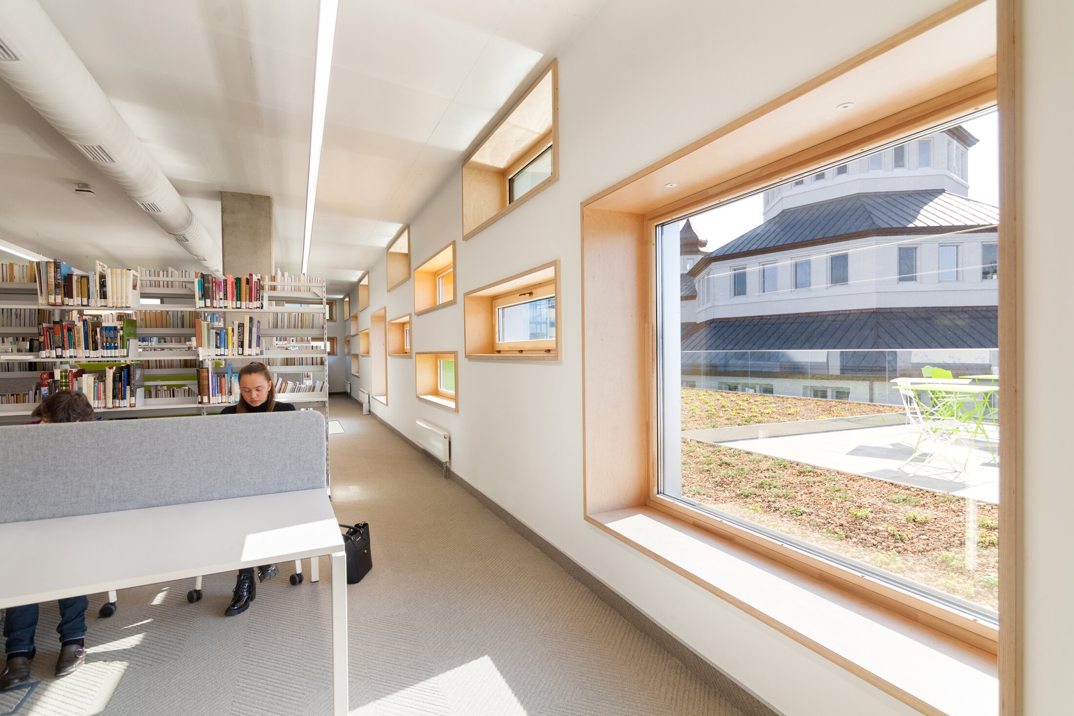 Project by Behnisch Architekturbuero, The Metropolitan Sheptytsky Center, Ukrainian Catholic University Lviv. Library study area with desks and bookshelves, small wood-framed windows. The view out of the window shows of a building and the terrace outside.