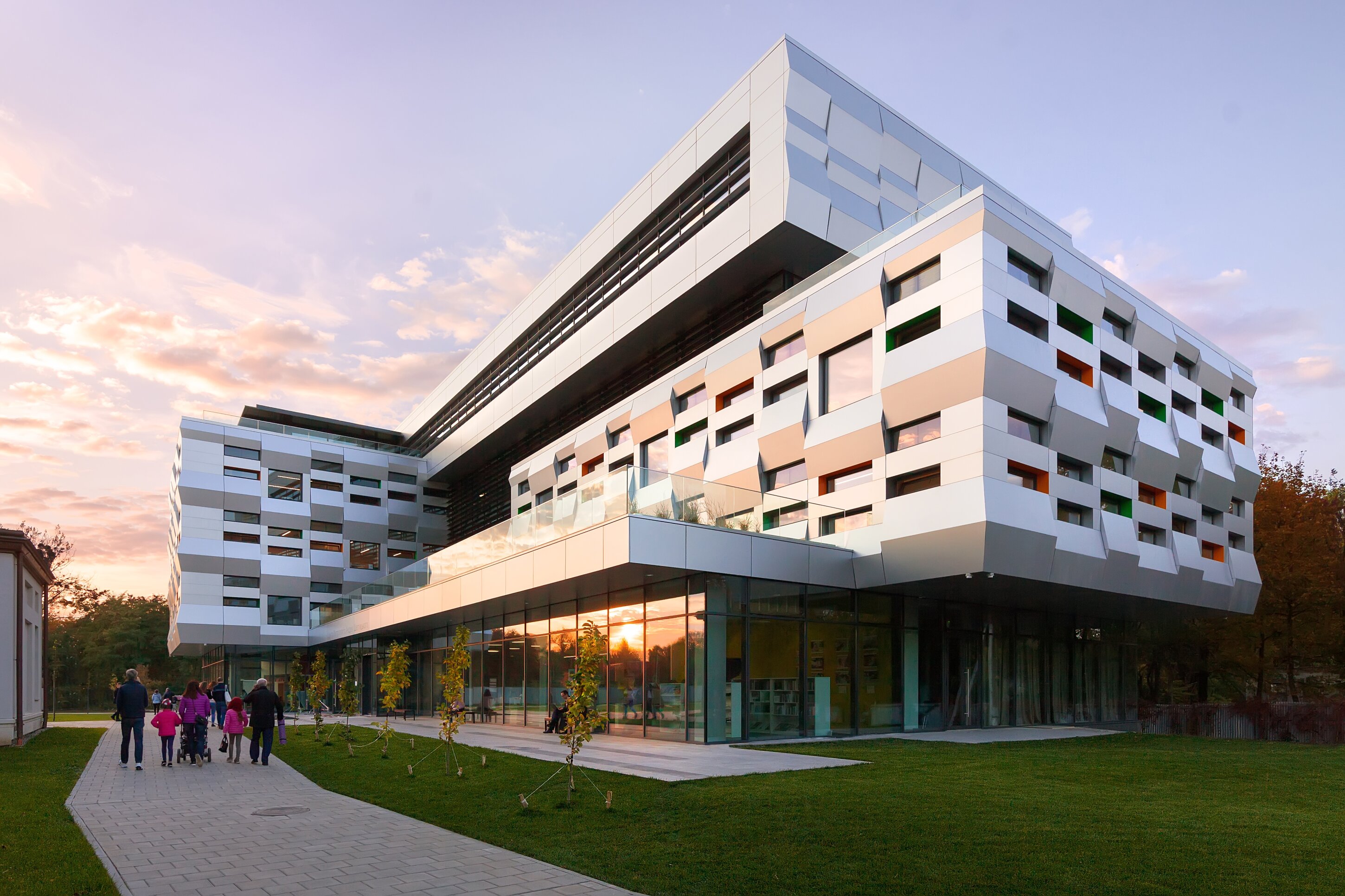 Project by Behnisch Architekturbuero, The Metropolitan Sheptytsky Center, Ukrainian Catholic University Lviv. Multi-story building with patterned facade and glass ground floor at sunset, with people walking along a path beside a lawn.