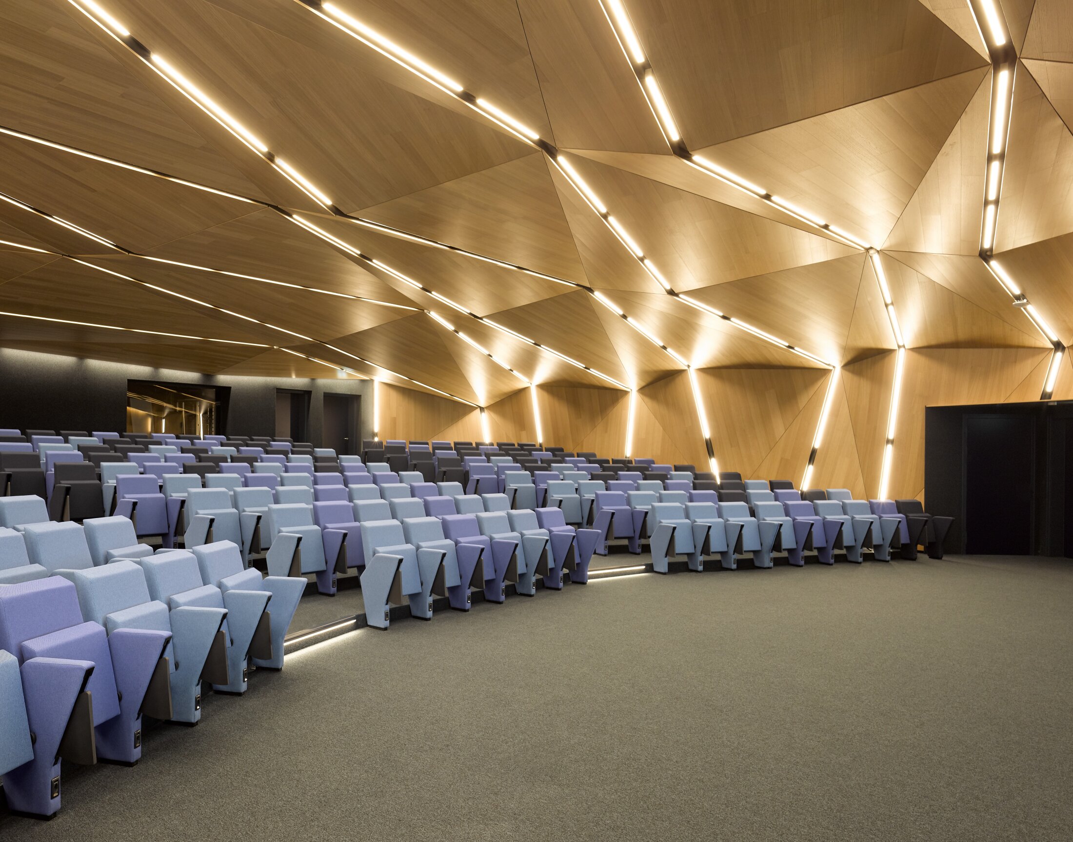 Project by Behnisch Architekturbuero, AGORA P&ocirc;le de recherche sur le cancer. Auditorium with tiered blue seats and a faceted wooden ceiling featuring integrated linear lighting panels.