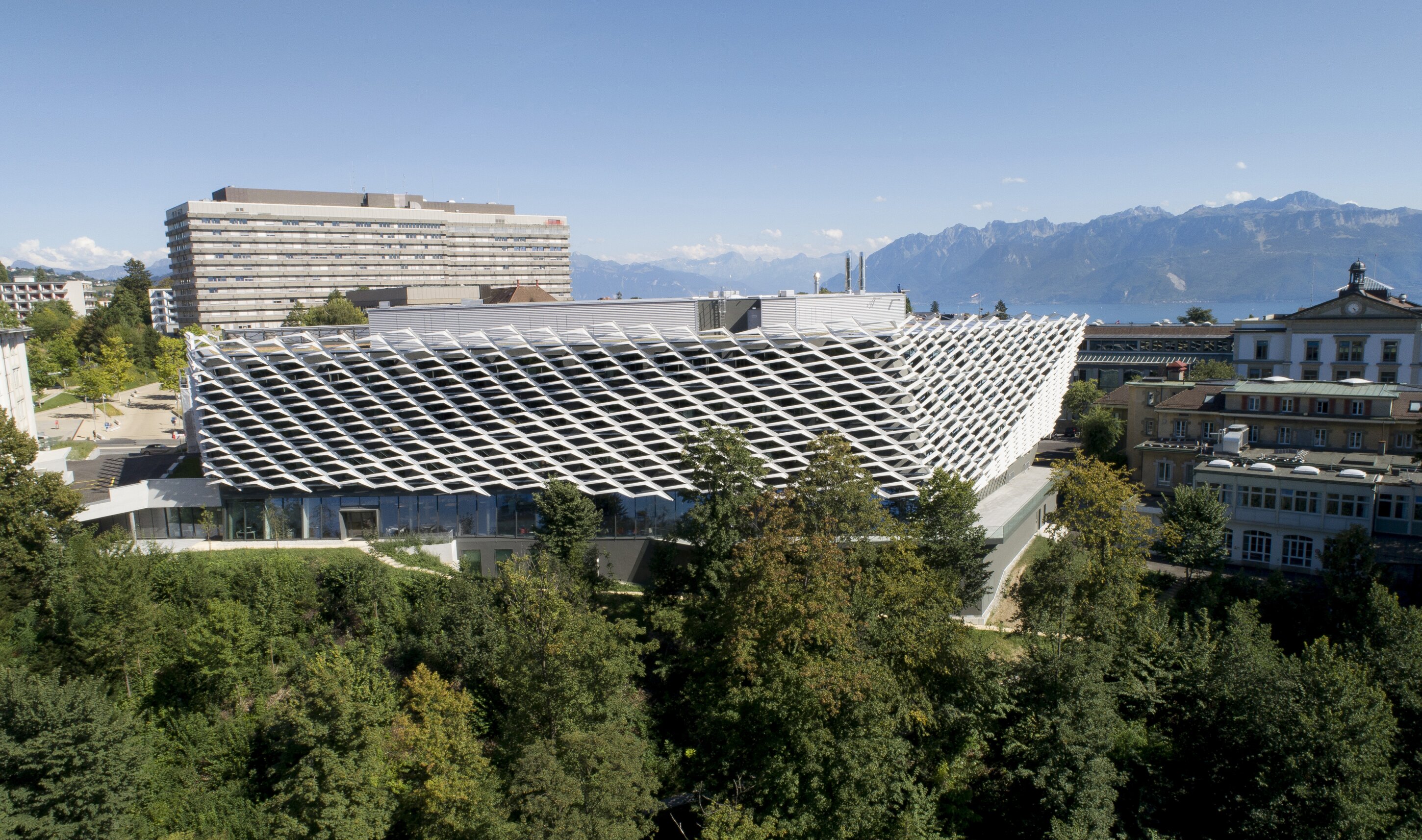 Project by Behnisch Architekturbuero, AGORA P&ocirc;le de recherche sur le cancer. Aerial view of a lakeside city with dense buildings. The large modern structure in the foreground, and mountains across the water under a clear sky.