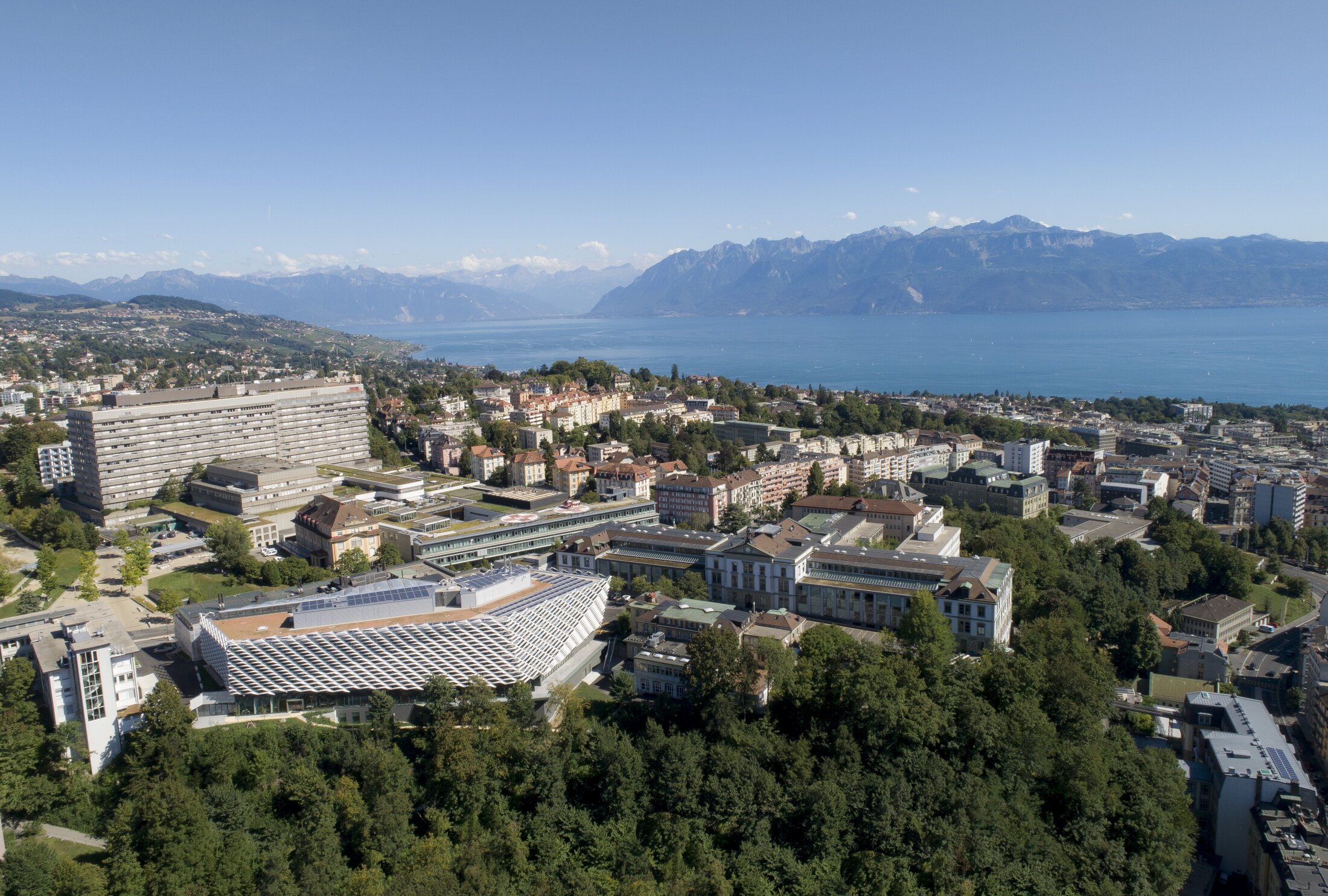 Project by Behnisch Architekturbuero, AGORA P&ocirc;le de recherche sur le cancer. Aerial view of a lakeside city with dense buildings. The large modern structure in the foreground, and mountains across the water under a clear sky.