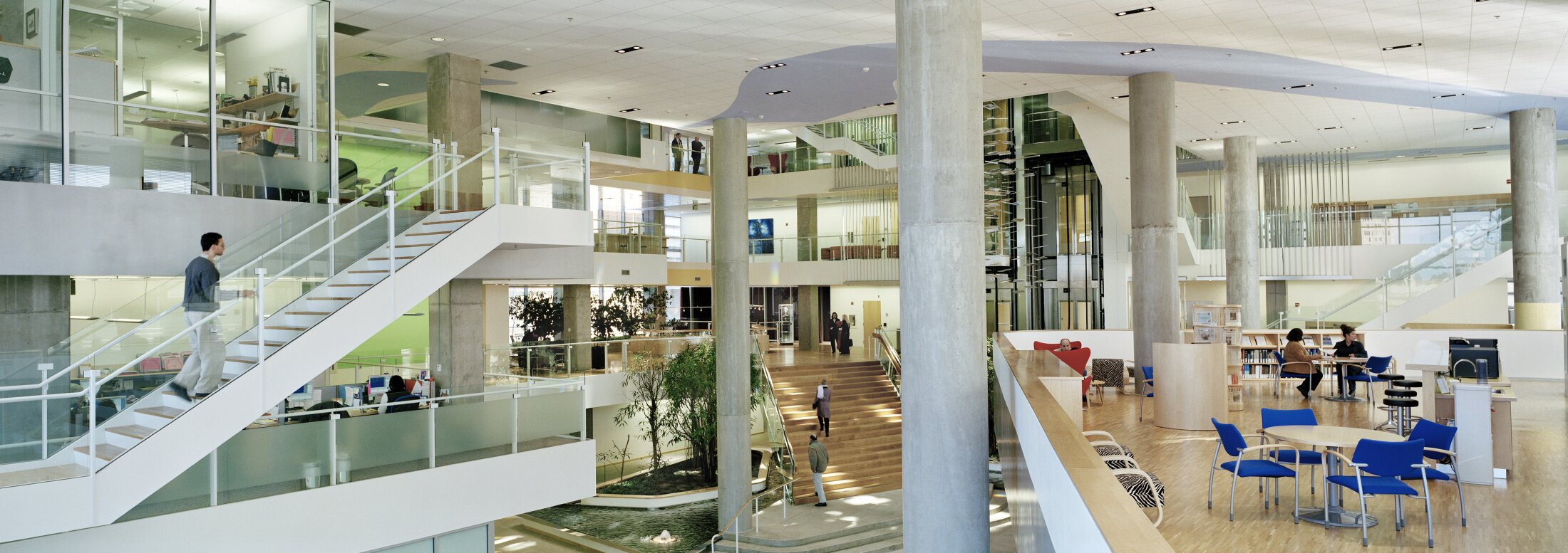 Project by Behnisch Architekturbuero, Genzyme Center. A large, multi-story atrium with stairs, glass railings, and columns. People walk around and linger in a seating area with tables and blue chairs.