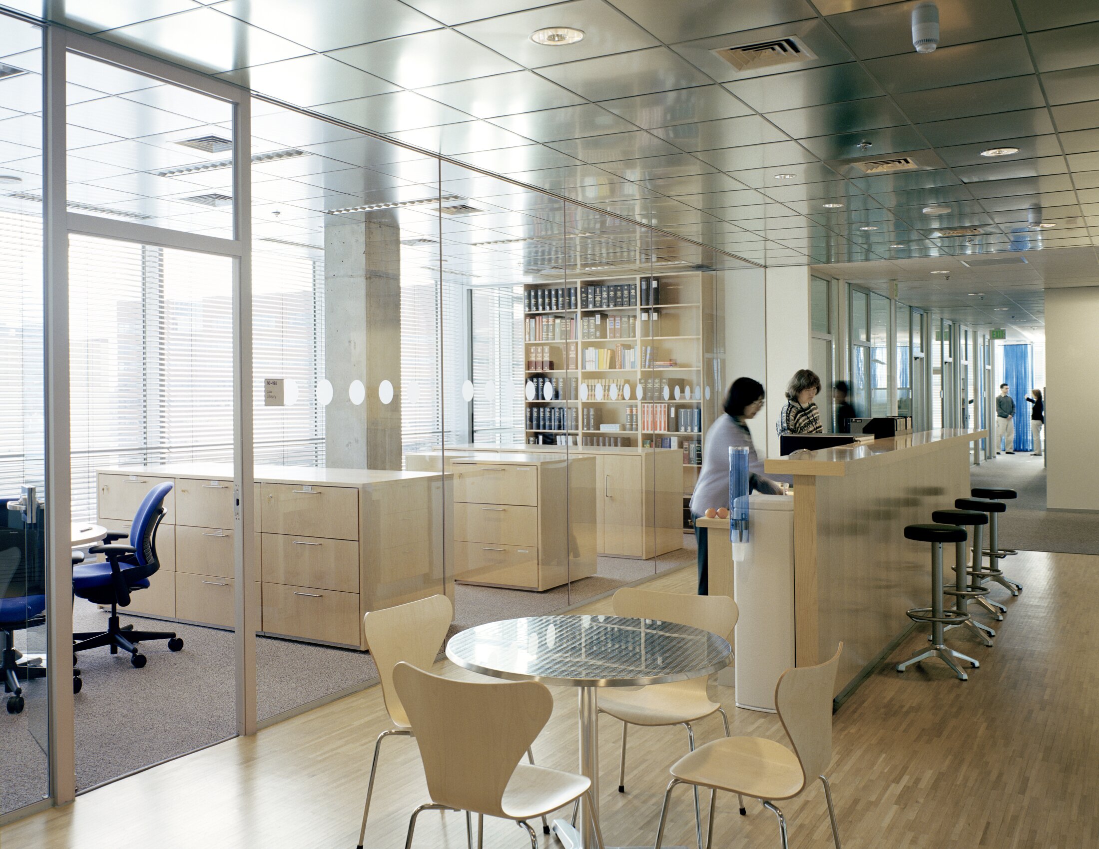 Project by Behnisch Architekturbuero, Genzyme Center. Office interior with glass partitions, desks and shelves, a counter with stools and a small table with chairs in the foreground. Two women are standing at the counter.