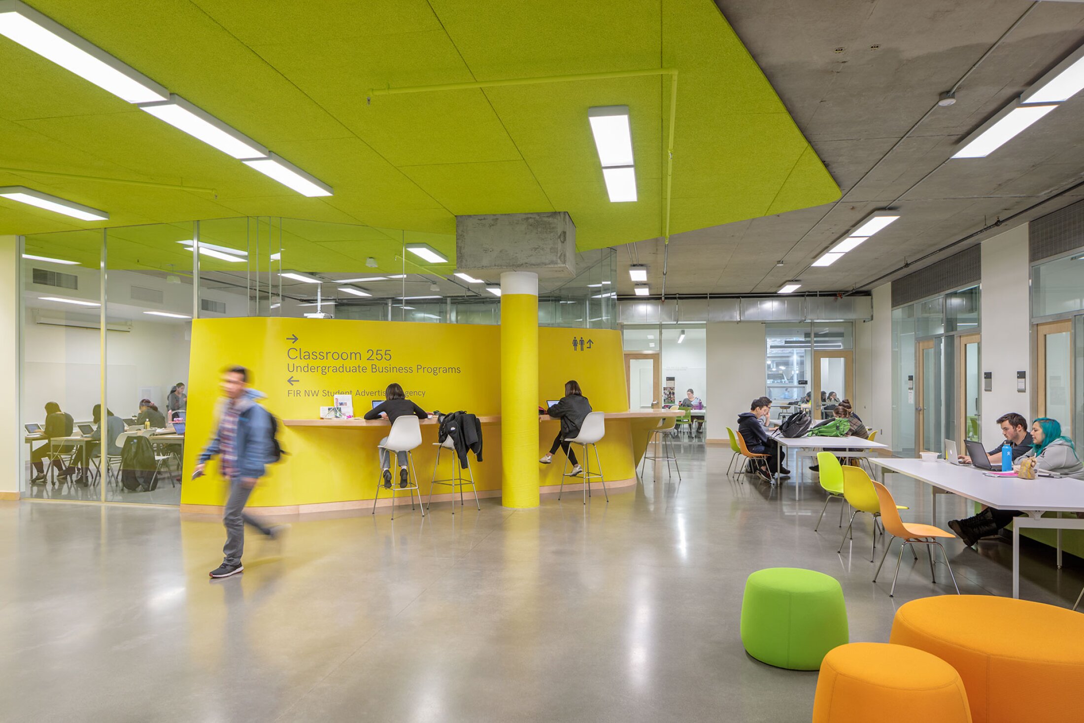 Project by Behnisch Architekturbuero, Karl Miller Center. Open study area with bright yellow walls and grenn acusticceiling. Students are working at tables and a central counter.