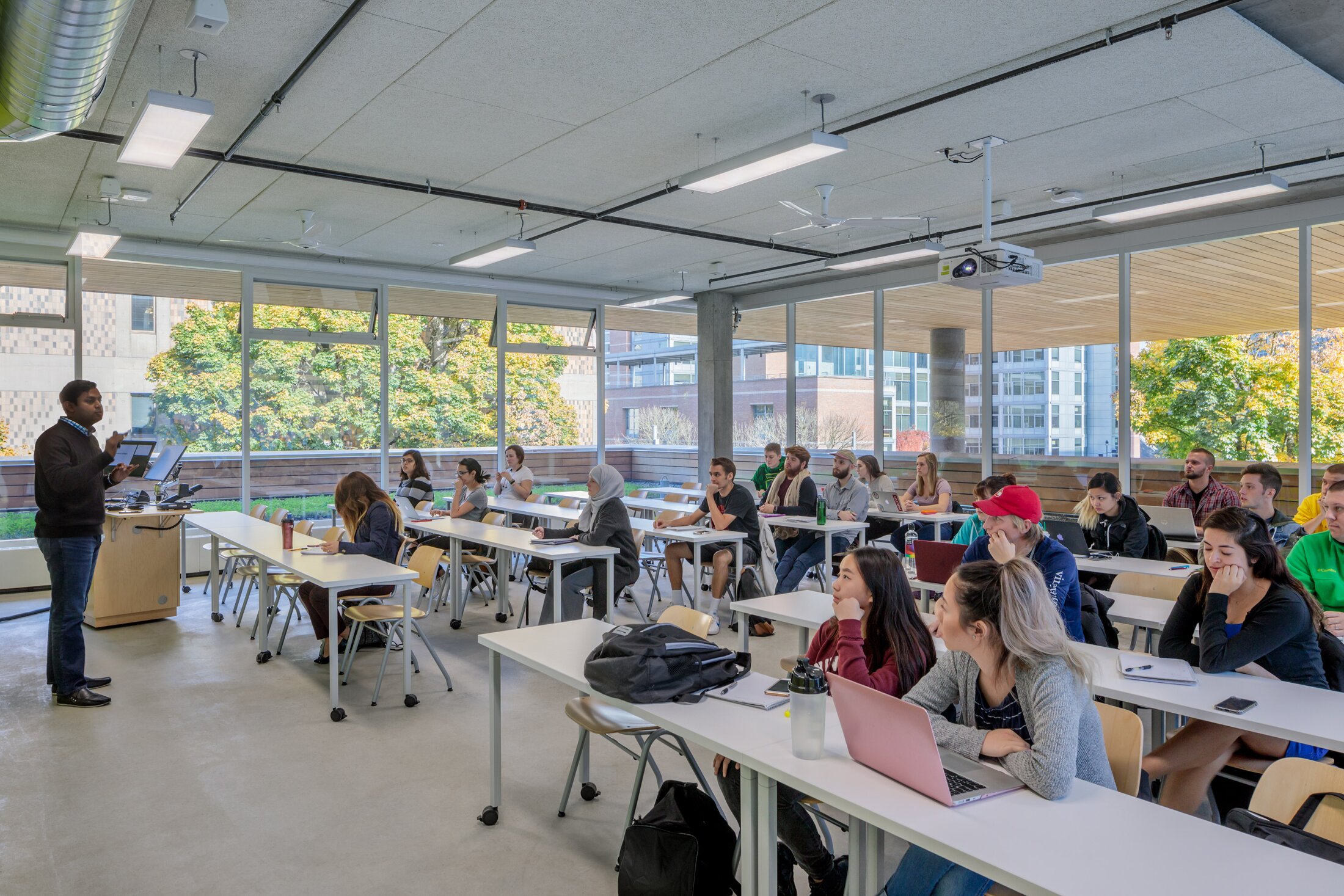 Project by Behnisch Architekturbuero, Karl Miller Center. Classroom with students seated at desks listening to an instructor speaking at the front near a lectern and screen.