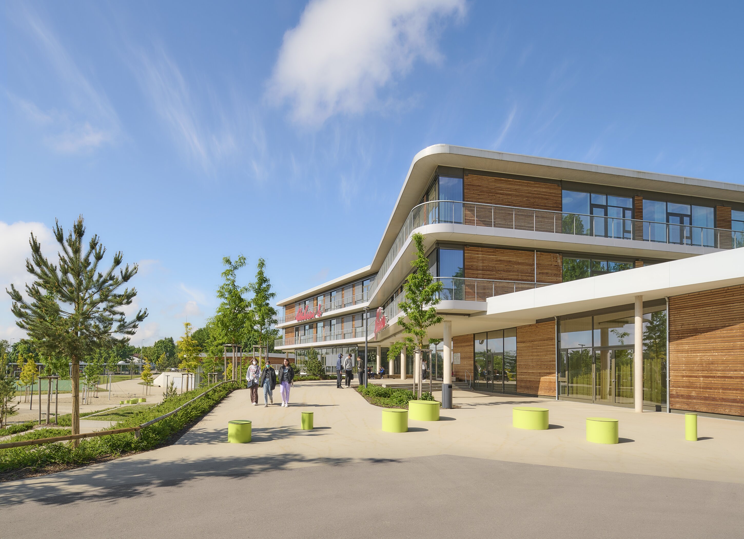 Project by Behnisch Architekturbuero, Anna-Pr&ouml;ll Secondary School and Sports Hall. Modern building with glass and wood facade, curved balconies, and a walkway with people and small green bollards under a blue sky.
