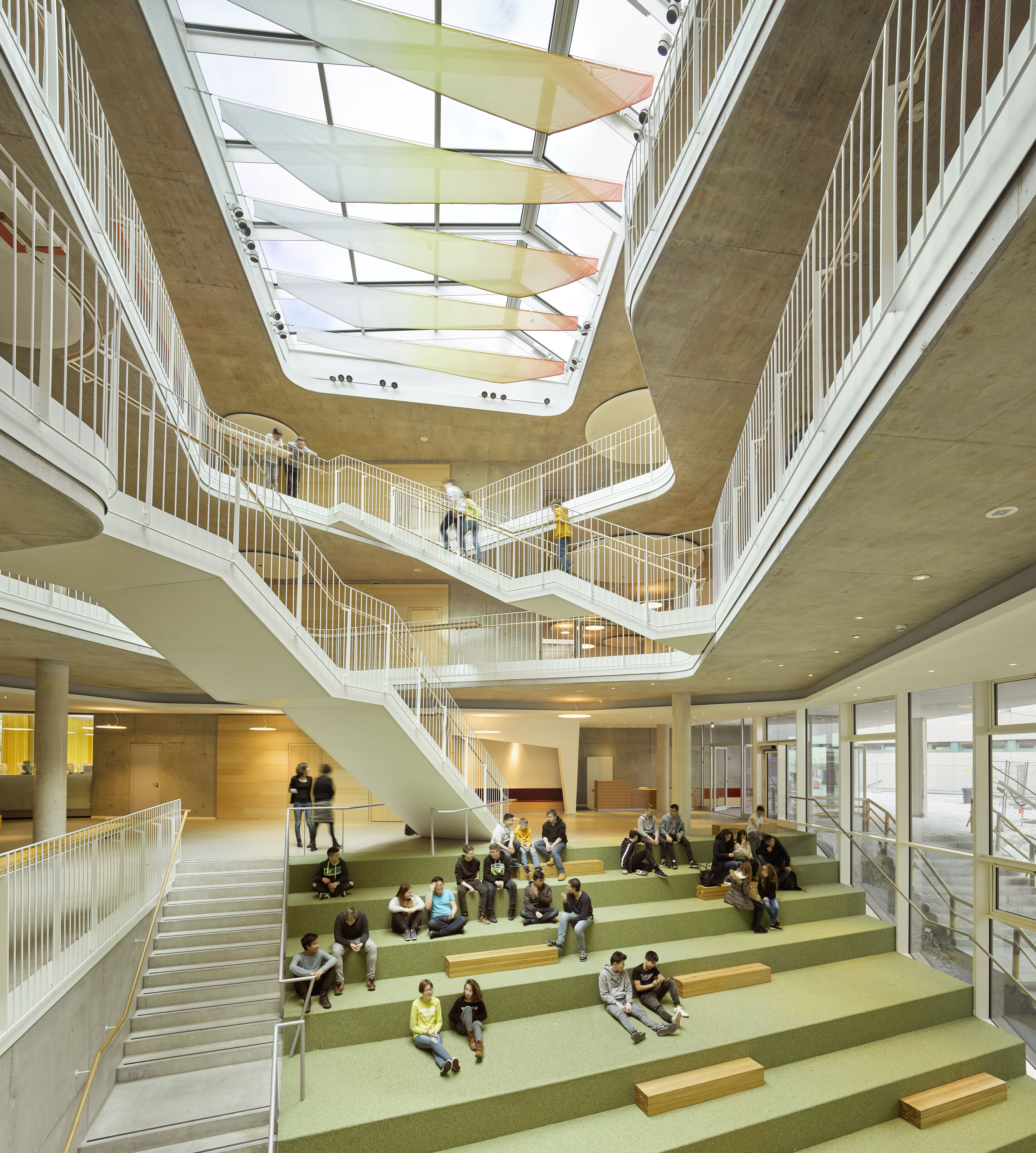 Project by Behnisch Architekturbuero, Anna-Pröll Secondary School and Sports Hall. Large school atrium with tiered seating, students sitting and talking, multiple staircases and walkways under a skylight ceiling.