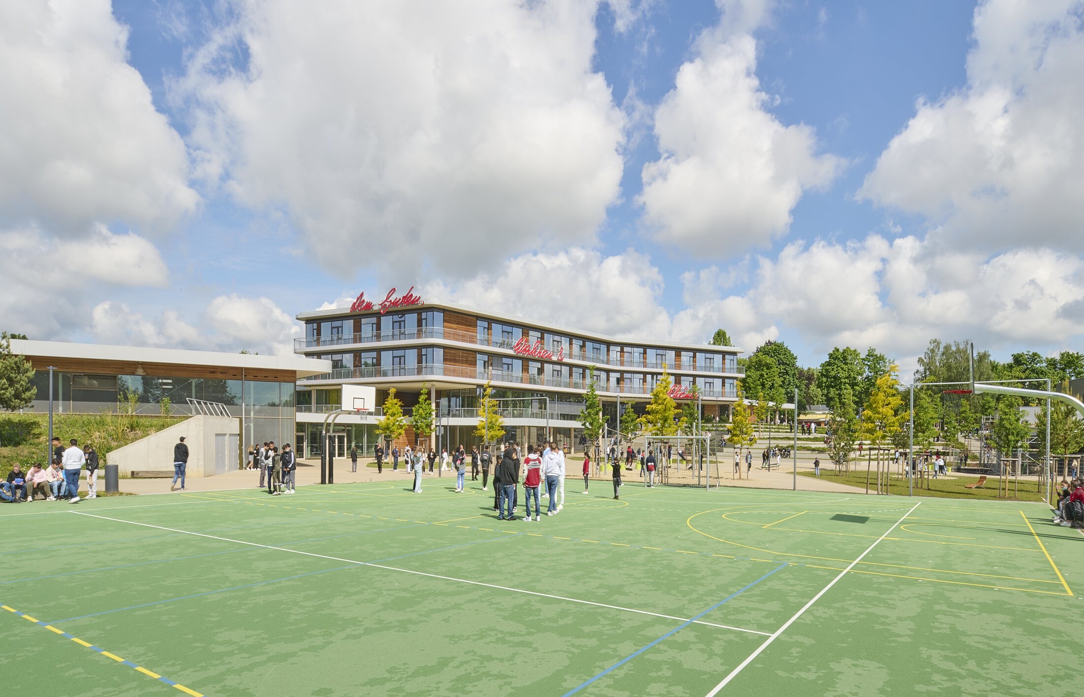 Project by Behnisch Architekturbuero, Anna-Pr&ouml;ll Secondary School and Sports Hall. School campus with students gathered on an outdoor sports court. In the background the Schoolbuilding, trees and a playground equipment nearby.