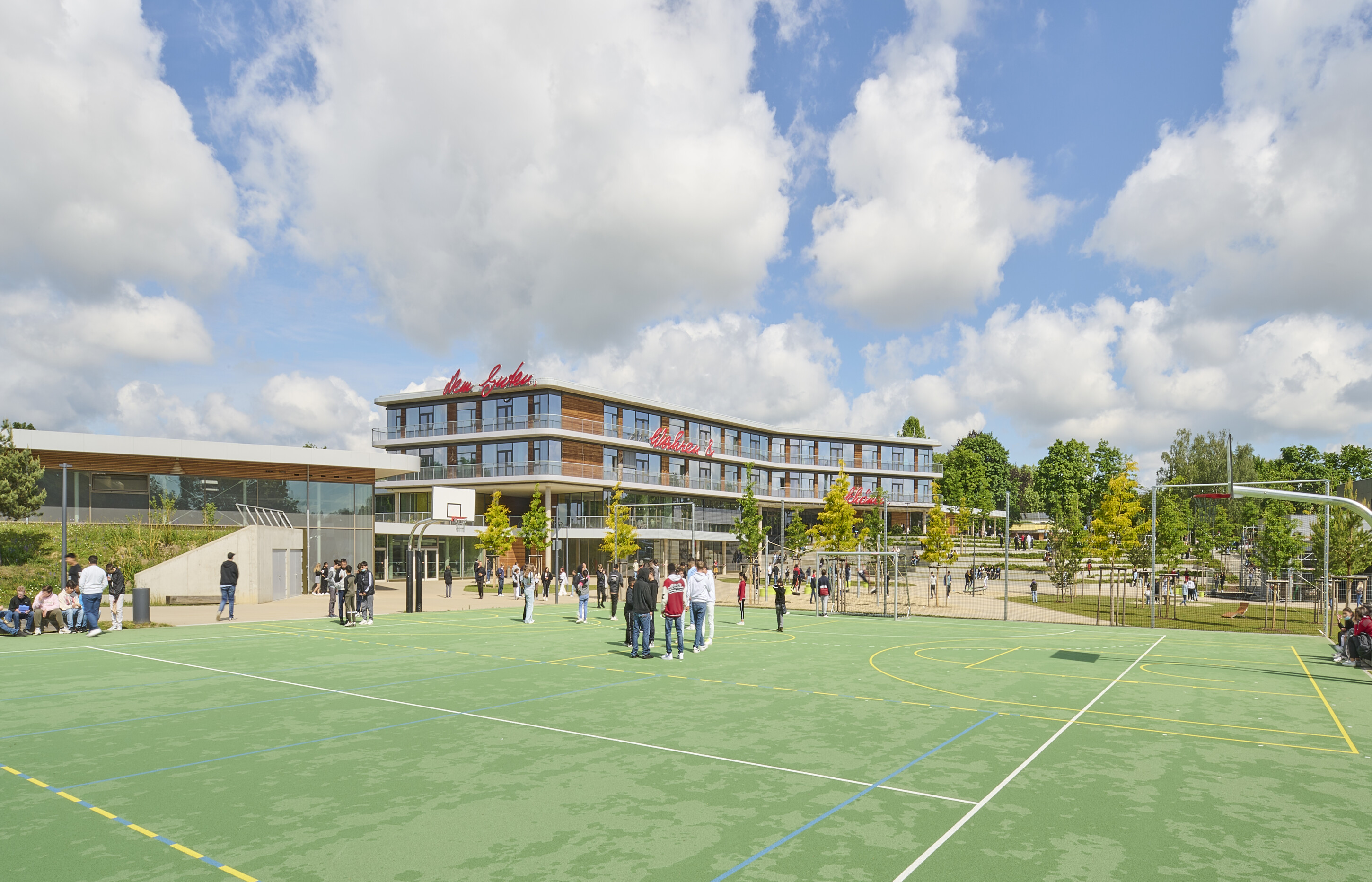 Project by Behnisch Architekturbuero, Anna-Pröll Secondary School and Sports Hall. School campus with students gathered on an outdoor sports court. In the background the Schoolbuilding, trees and a playground equipment nearby.