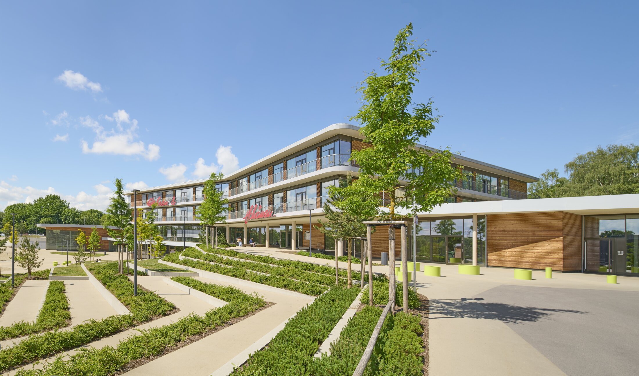 Project by Behnisch Architekturbuero, Anna-Pr&ouml;ll Secondary School and Sports Hall. Modern school building with glass and wood facade, landscaped terraces and paths, trees and open entrance area under blue sky.