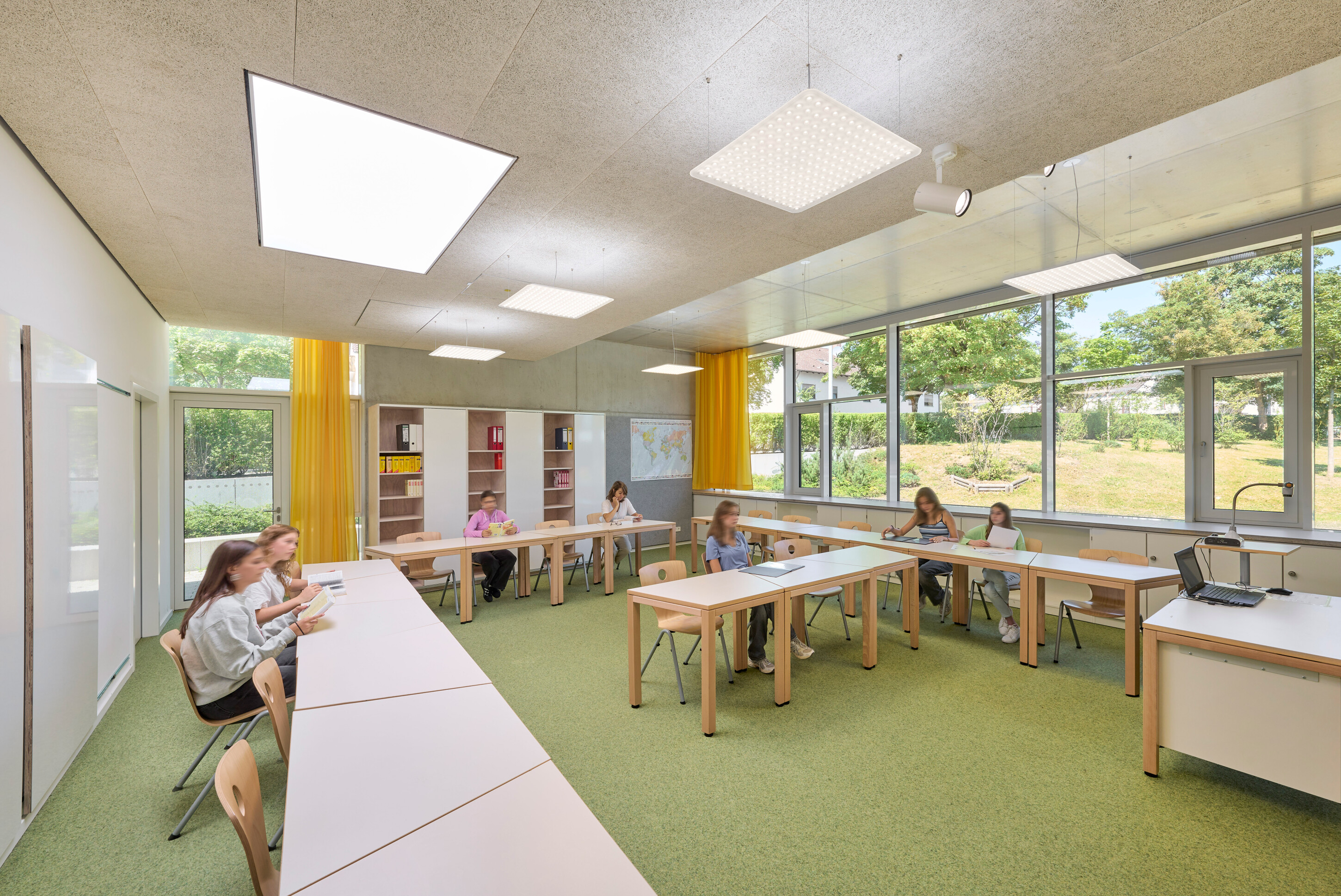 Project by Behnisch Architekturbuero, Anna-Pröll Secondary School and Sports Hall. Classroom with students seated at tables in a U-shape, reading and writing. The room has large windows, shelves, and yellow curtains. The floor is in a light green.