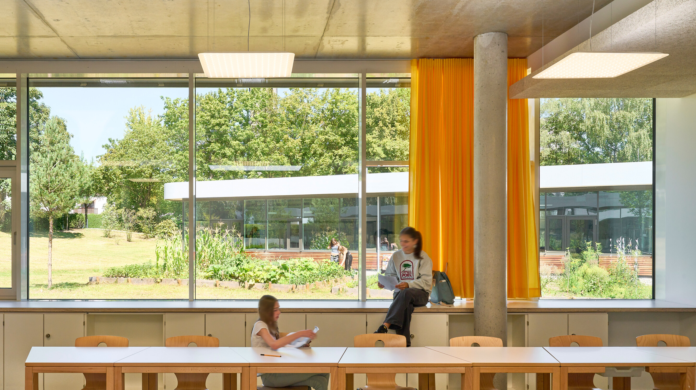 Project by Behnisch Architekturbuero, Anna-Pröll Secondary School and Sports Hall. Classroom with large windows and yellow curtains, one teacher and one student reading. The tables and chairs are in the foreground, the garden is visible outside.