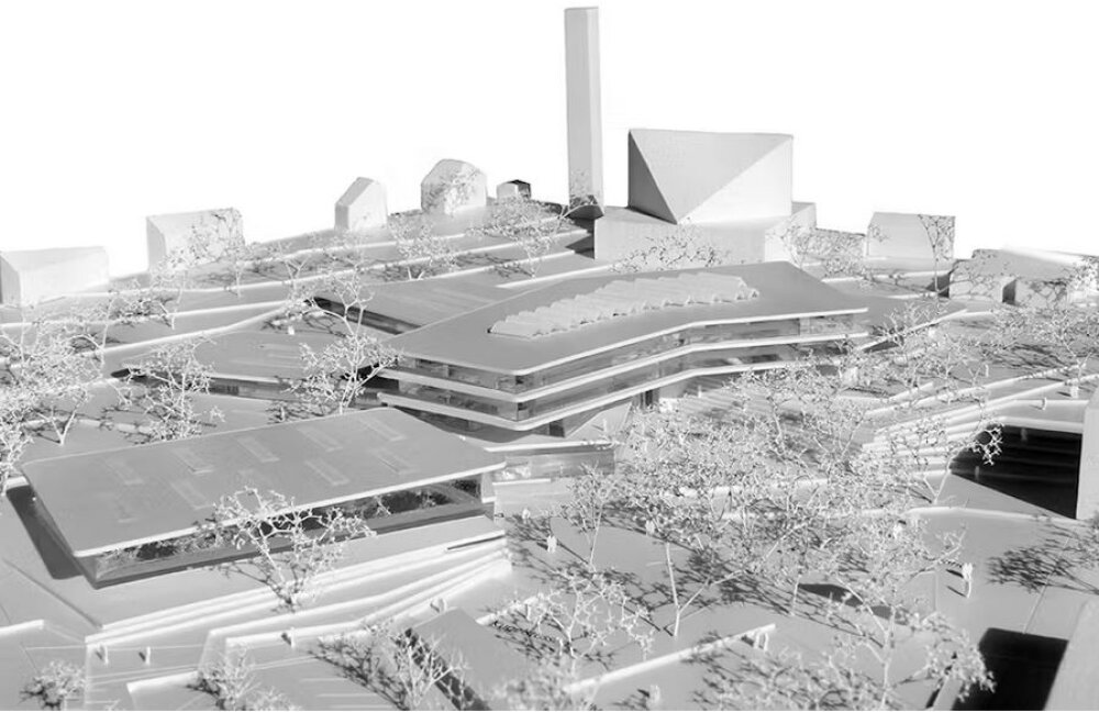 Project by Behnisch Architekturbuero, Anna-Pr&ouml;ll Secondary School and Sports Hall. Architectural model showing a multi-level complex with curved buildings, surrounding trees, and nearby streets and structures.