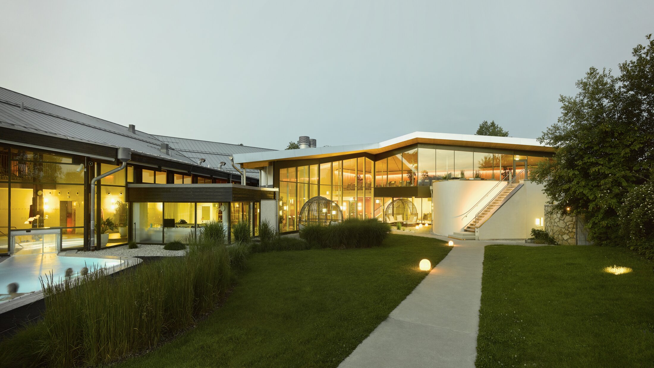Project by Behnisch Architekturbuero, Thermal Spa Bad Aibling. Modern spa complex at dusk with glass facades, lit interiors, lawn, pathway lights, and outdoor pool visible on the left.