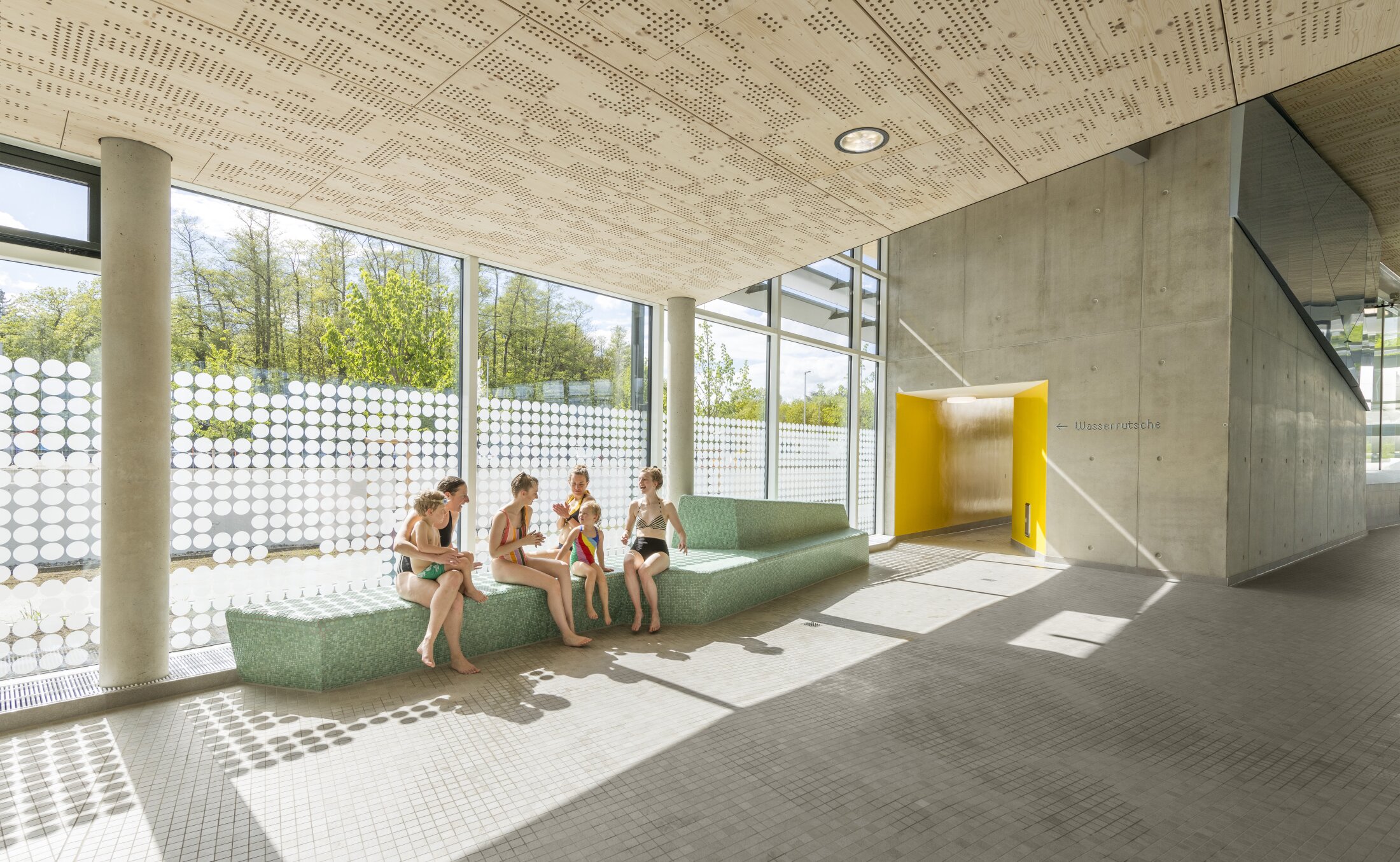 Parents and children in swimwear sit on a bench in a bright indoor swimming pool area in front of large windows. Concrete walls and a yellow entrance to the water slides are visible in the background.