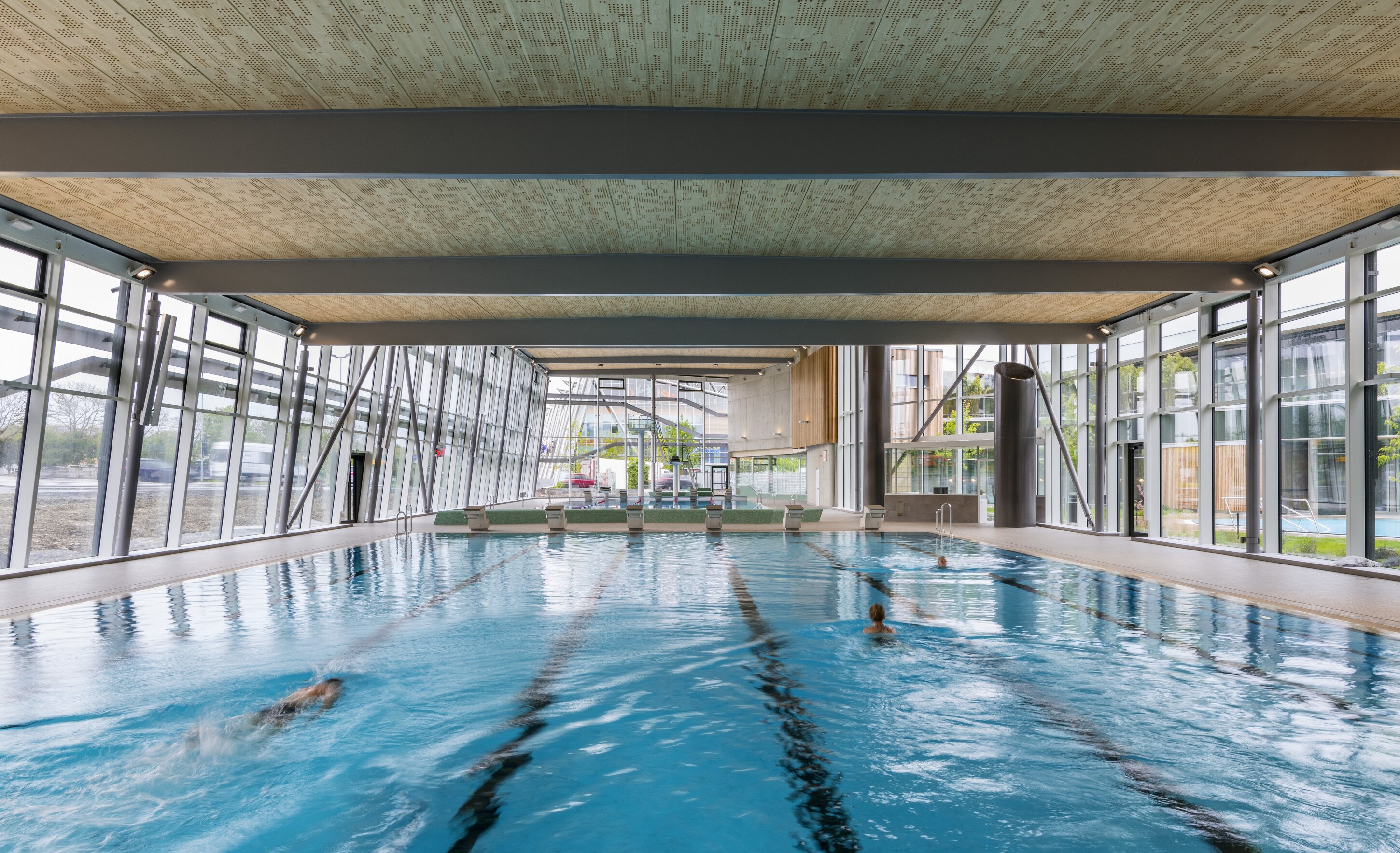 Project by Behnisch Architekturbuero, Sportbad Friedrichshafen. Indoor lap pool with swimmers, lane markings, and large glass walls under a timber ceiling in a modern aquatic center.