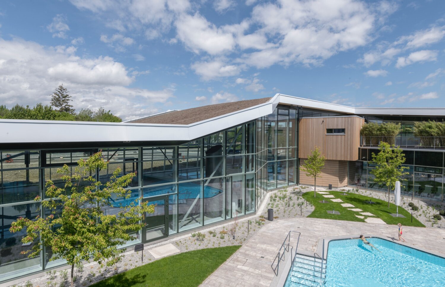 Project by Behnisch Architekturbuero, Sportbad Friedrichshafen. Courtyard with glass buildings around a pool, landscaped garden with trees and umbrellas, and terraces under a partly cloudy sky. People are swimming in the pool.