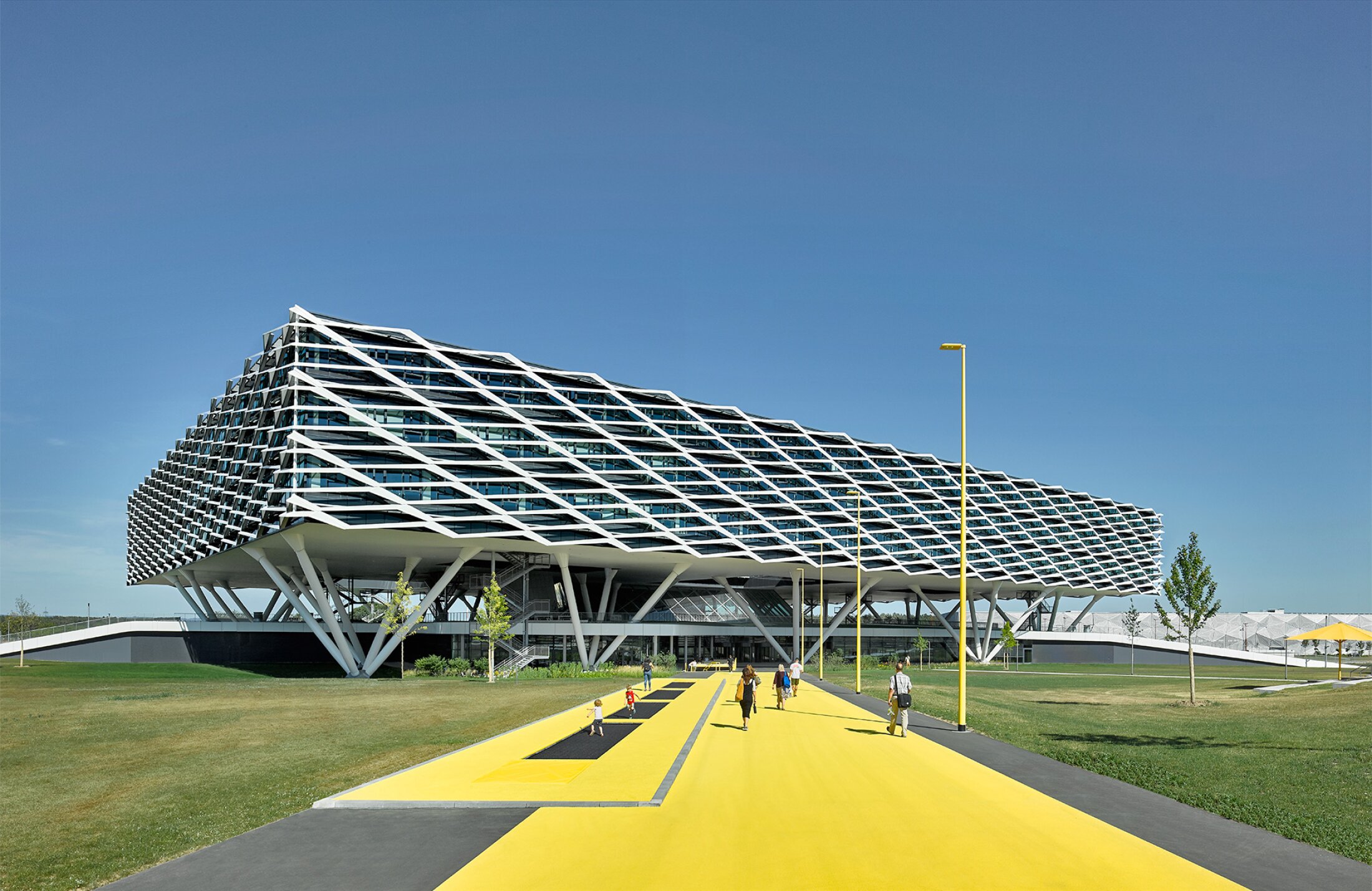 Project by Behnisch Architekturbuero, adidas ARENA. A large, modern building with a sloping, grid-like facade features it is a specially designed sun protection and shading system. The main building is elevated and stands on stilts. People walk along a wide yellow path leading to the entrance.