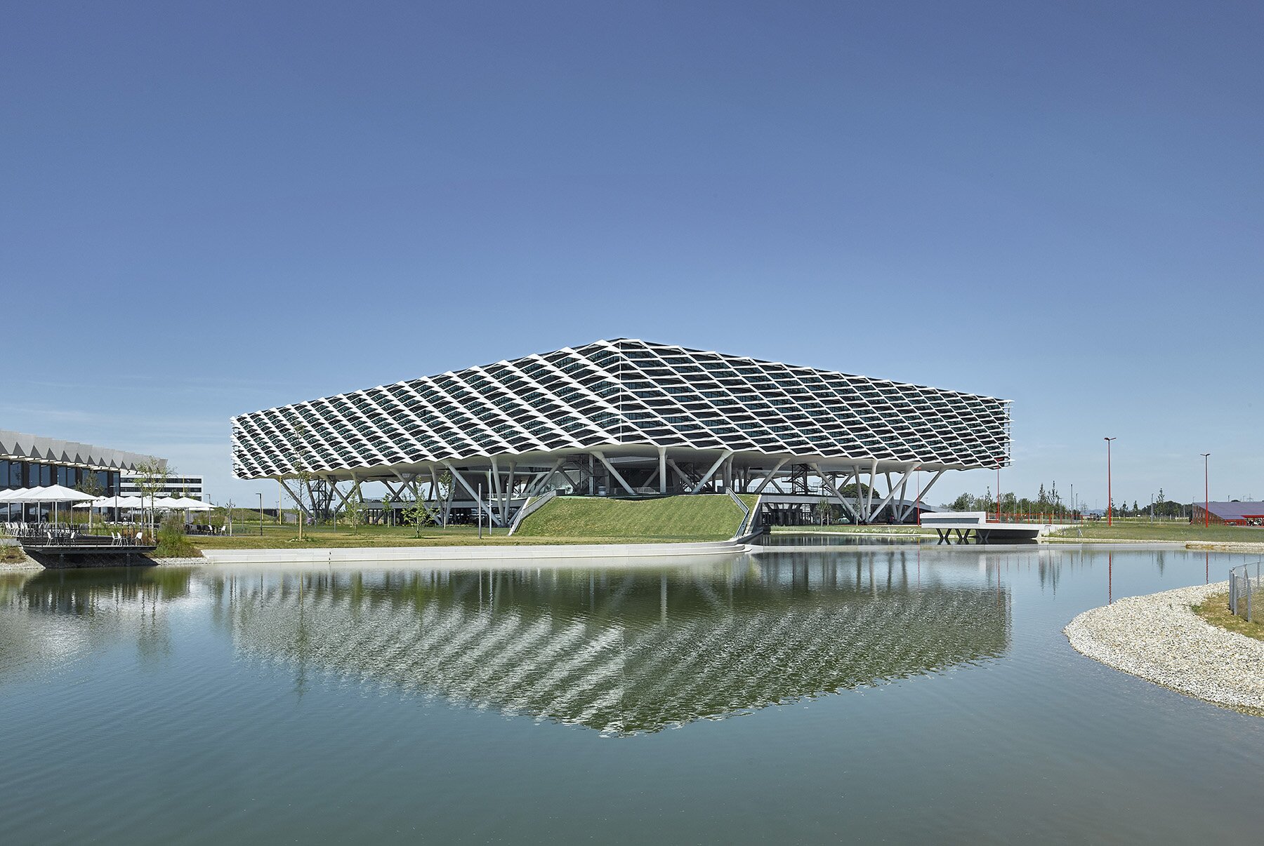Project by Behnisch Architekturbuero, adidas ARENA. A large, modern building with a sloping, grid-like facade features it is a specially designed sun protection and shading system. It is reflecting in the surface under a clear sky.